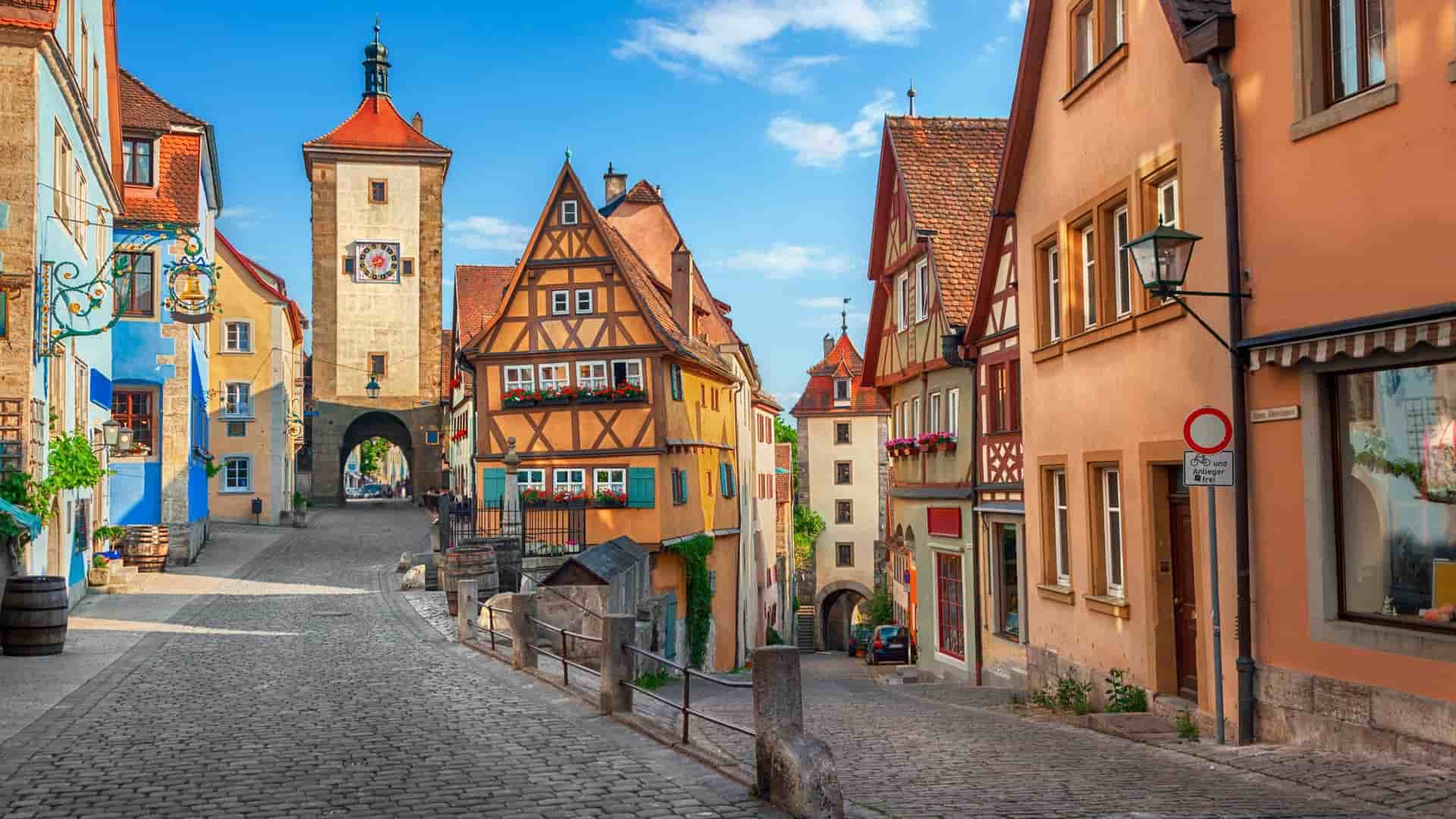 A charming view of the iconic Plönlein intersection in Rothenburg, Germany, featuring a cobblestone street, colorful half-timbered houses, and the Siebers Tower under a bright blue sky.