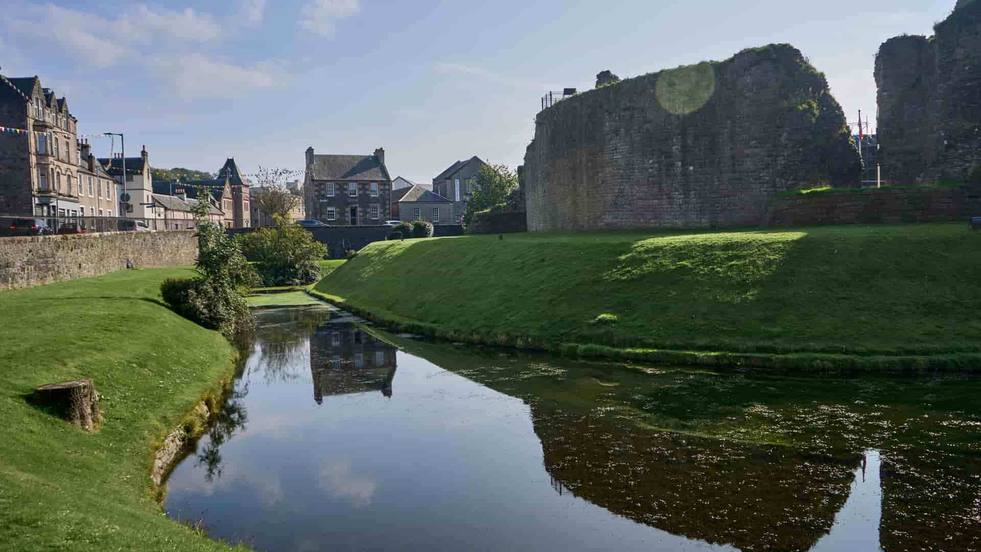 A picturesque view of the ruins of the medieval Rothesay Castle on the Isle of Bute, Scotland, with a tranquil moat and green lawn in front of historic stone houses.