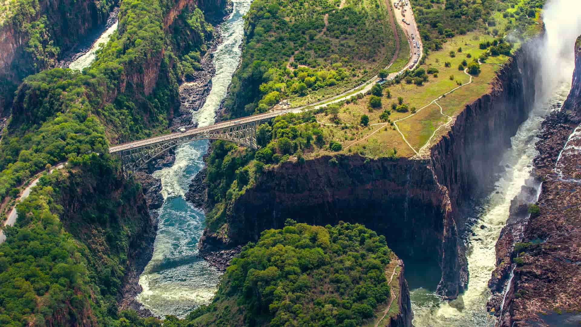 An aerial view of Victoria Falls, showcasing the magnificent waterfall and the Victoria Falls Bridge spanning the Zambezi River, with a train visible on the bridge and lush green gorges surrounding the area.