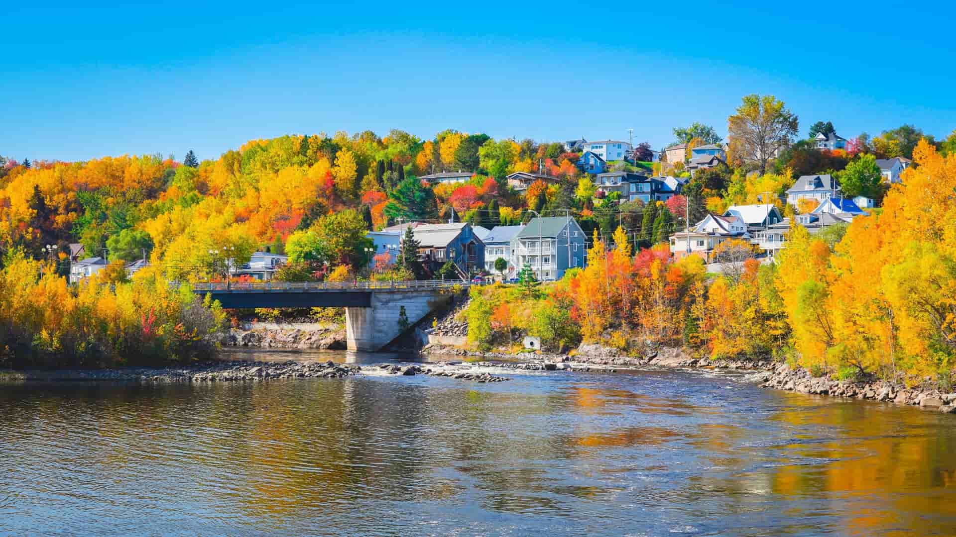 A beautiful autumn landscape in Saguenay with a river flowing under a bridge, surrounded by hillsides adorned with houses and vibrant yellow, orange, and red fall foliage.