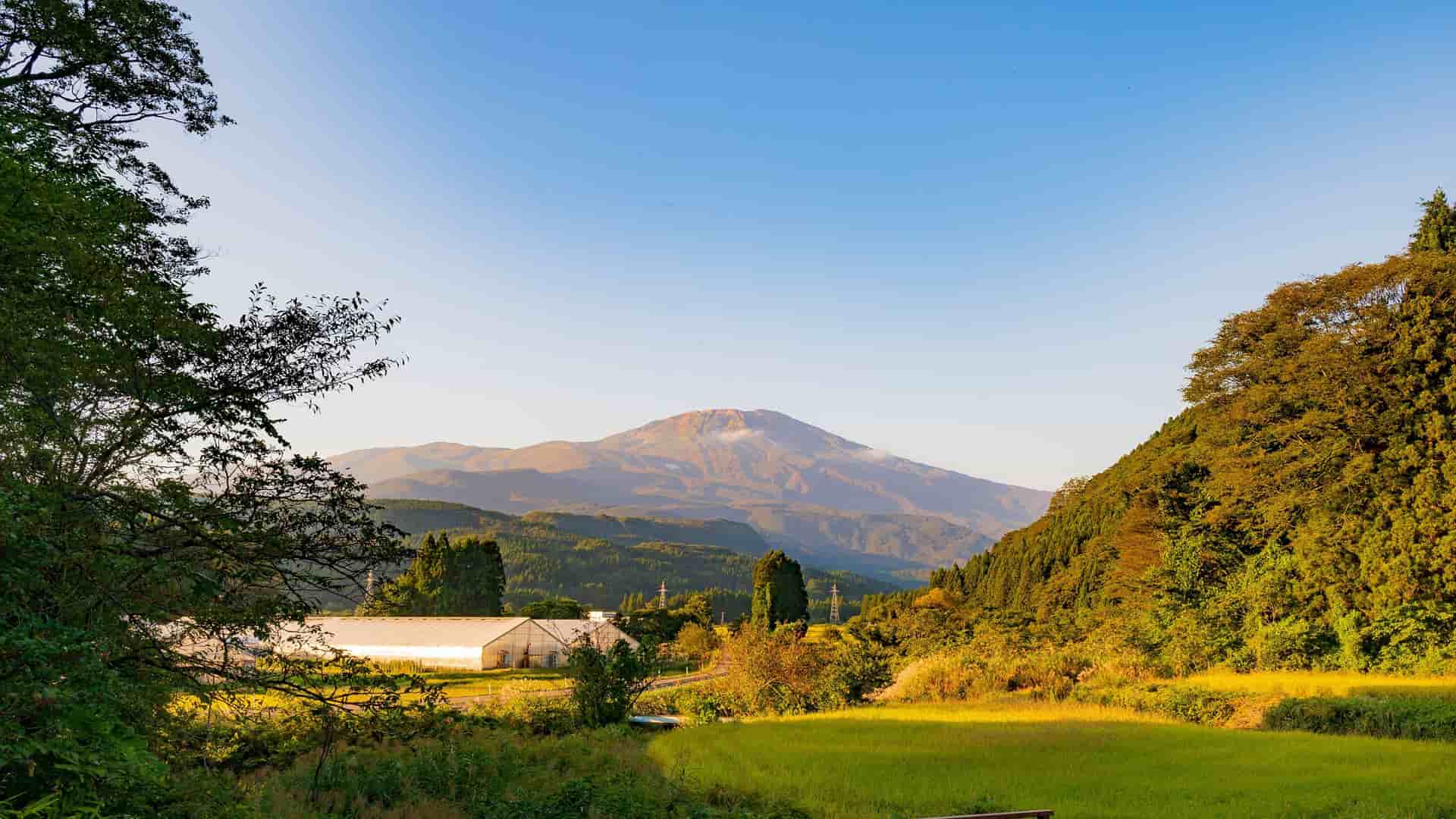 A beautiful landscape of rural Sakata, Japan, with green fields, a farmhouse, and lush forests in the foreground, and the majestic, distant Mount Chokai under a clear blue sky.