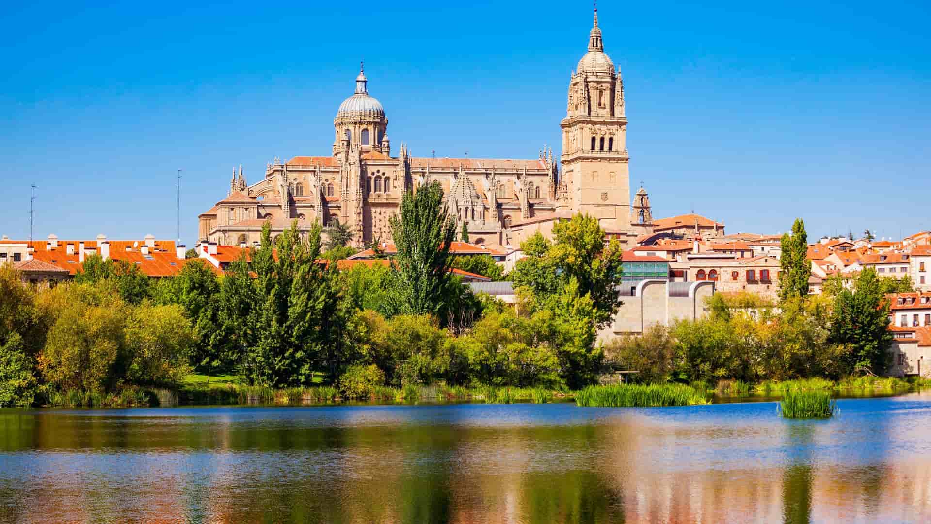 A beautiful panoramic view of the historic Salamanca Cathedral, with its twin towers and dome, majestically rising above the cityscape and reflecting on the calm waters of the Tormes River under a vibrant blue sky.