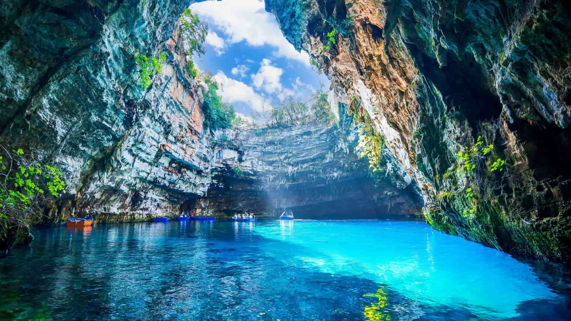 A mesmerizing view of Melissani Cave Lake in Sami, Greece, with its striking turquoise-blue water and a large opening in the cave ceiling that lets in a beam of natural sunlight.