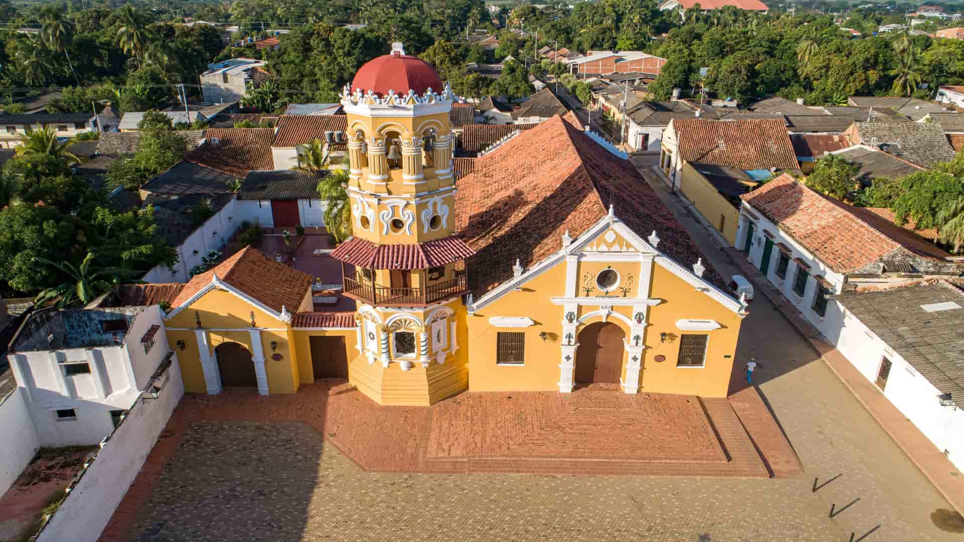 An aerial view of a vibrant yellow colonial church with a red-tiled roof and bell tower, the main landmark of the historic town of Santa Bárbara de Pinto in Colombia.