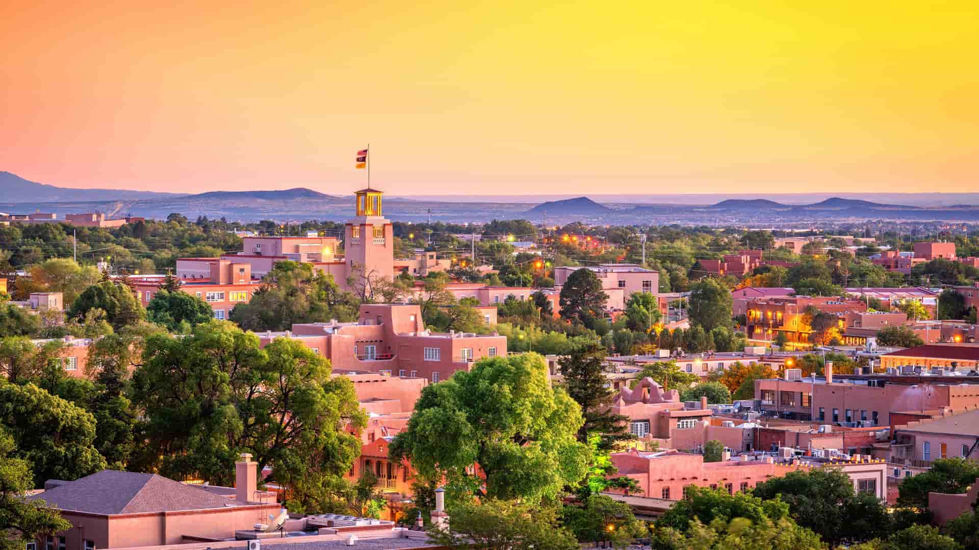 "A panoramic sunset view of the Santa Fe, New Mexico skyline with a yellow-orange sky, showing the distinctive pueblo-style architecture and surrounding desert landscape."