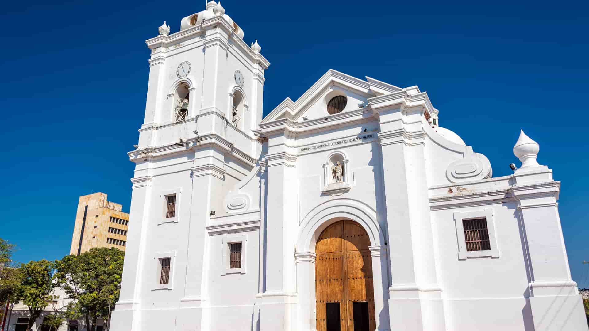 A white colonial-style cathedral with a bell tower and wooden doors is visible under a clear blue sky, likely in Santa Marta, Colombia.