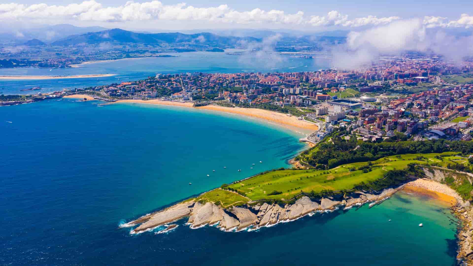 An aerial panoramic view of the beautiful coastal city of Santander, Spain, showcasing a long sandy beach, the Magdalena Peninsula with a lush golf course, and the sparkling blue bay.