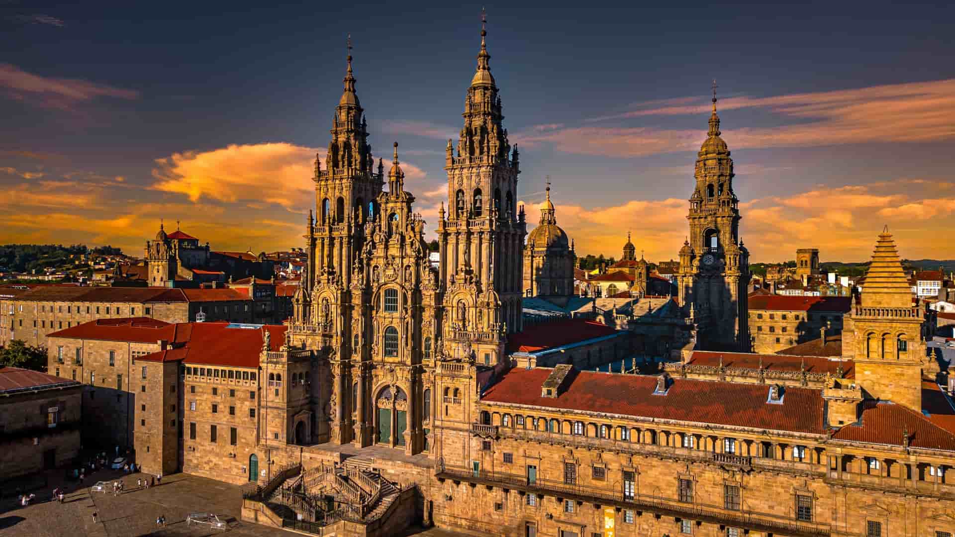 A beautiful sunset view of the Santiago de Compostela Cathedral, a stunning Romanesque and Baroque landmark with intricate carvings and towers, with the historic city and a dramatic, cloudy sky in the background.