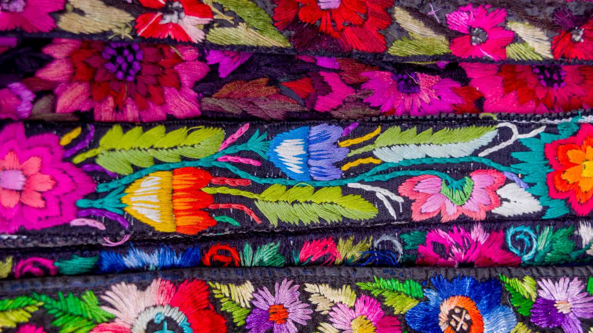A close-up of vibrant, traditional Mayan textiles from Santo Tomás de Castilla, Guatemala, featuring intricate, colorful hand-embroidered floral patterns on a dark fabric.
