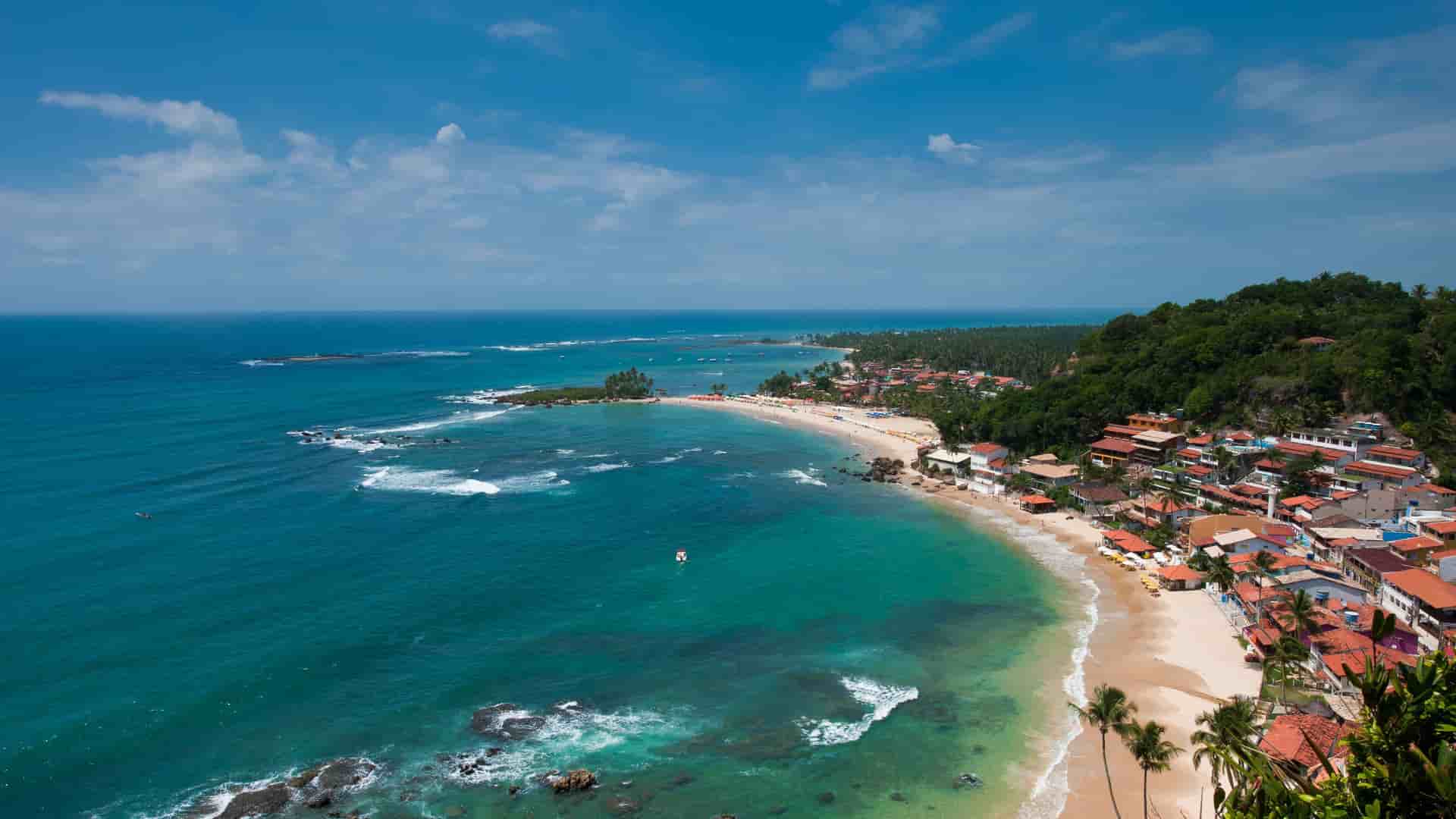 A scenic aerial shot of the tropical Morro de Sao Paulo beach in Brazil, showing the turquoise Atlantic Ocean, a long stretch of golden sand, and a village with red-roofed houses nestled against a lush green hill.