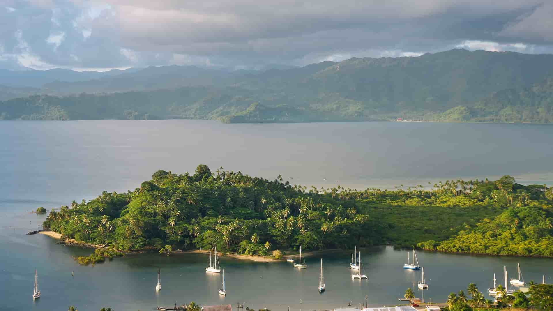 A panoramic view of Savusavu, Vanua Levu, Fiji, with a tranquil bay filled with sailboats, a lush green peninsula in the foreground, and mountainous terrain in the background.