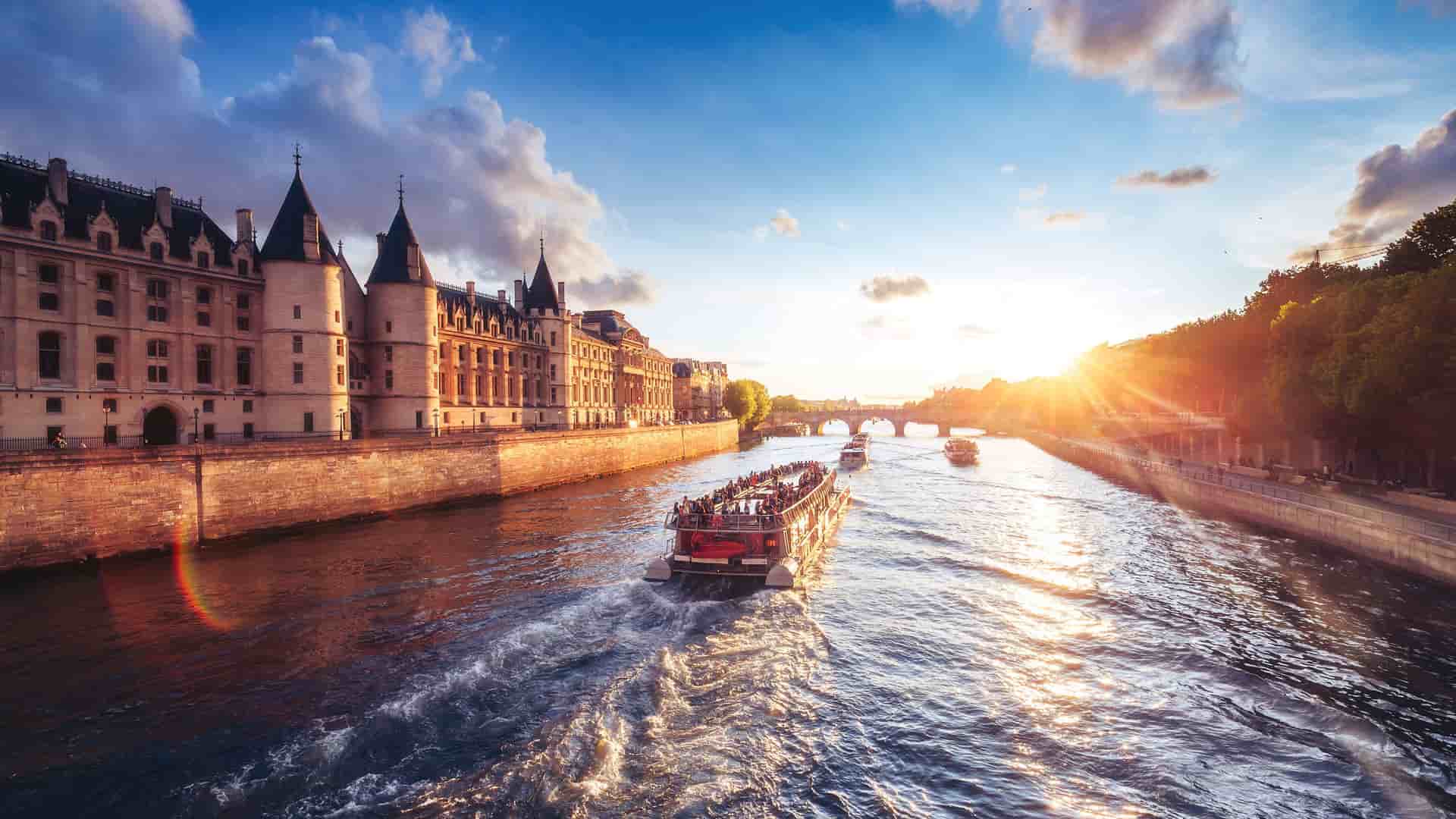 A vibrant sunset view of a river cruise boat on the Seine River in Paris, with the historic Conciergerie building and the Pont au Change bridge in the background.