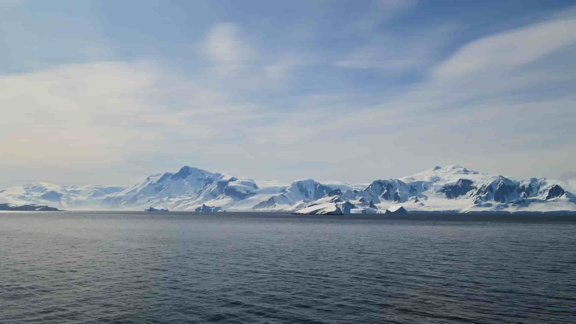 A panoramic view of the vast, icy landscape of Antarctica, with massive snow-covered mountains, glaciers, and icebergs floating in the deep blue waters of the Schollaert Channel.