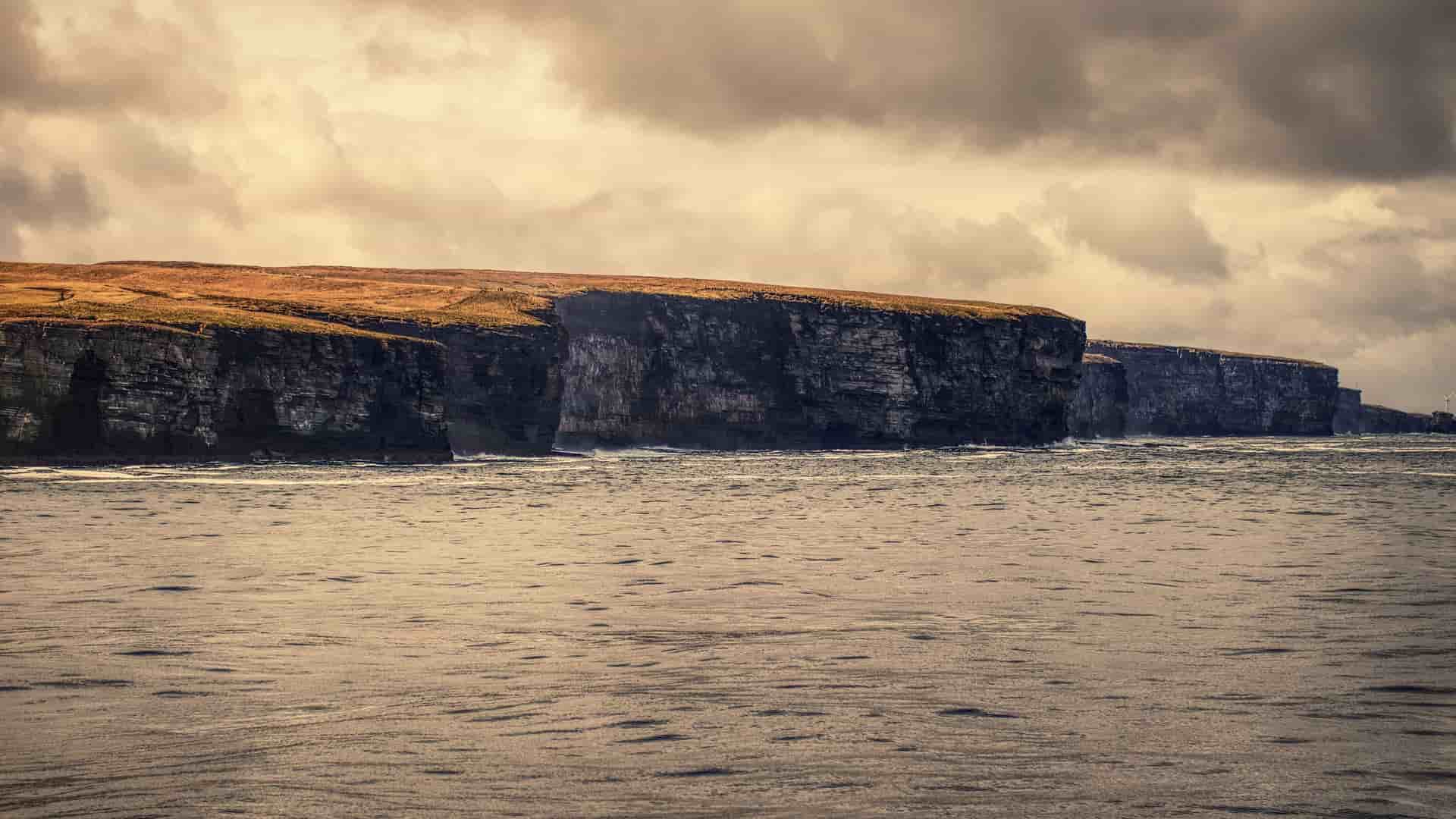 A scenic view of the dramatic, high sea cliffs on the coast of Scrabster, Scotland, with the vast, dark waters of the Pentland Firth in the foreground and a moody sky above.