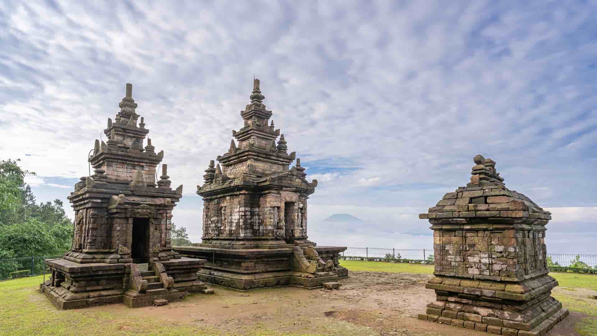 A stunning wide-angle view of the Gedong Songo temple complex, a collection of medieval Hindu temples on a misty hillside, with a mountain in the background in Semarang, Java, Indonesia.