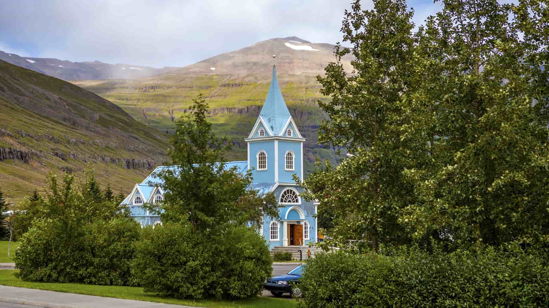 A scenic shot of the iconic blue church, Seyðisfjarðarkirkja, in Seyðisfjörður, Iceland, with a green mountain backdrop and lush trees in the foreground.