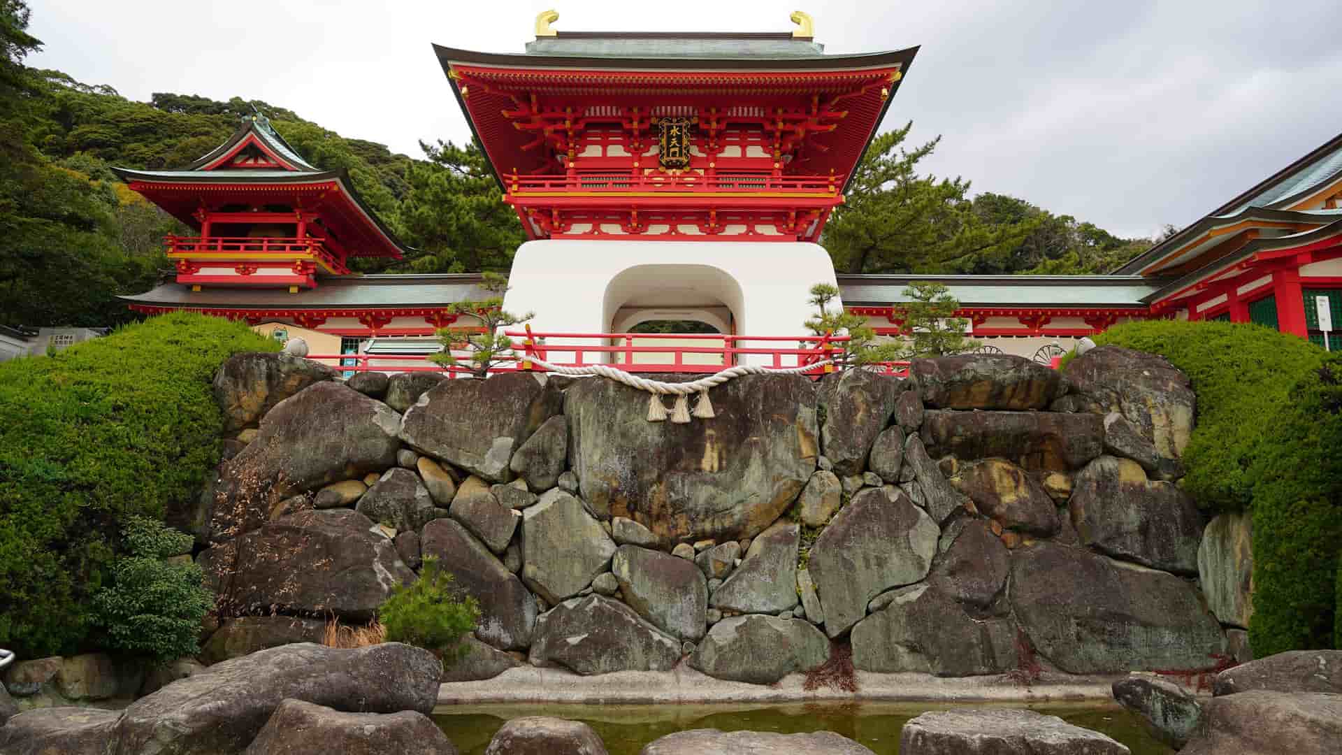 A stunning view of the Akama Shrine in Shimonoseki, Japan, with its vibrant red and white buildings and an intricate rope and paper ornament over a tranquil pool of water.