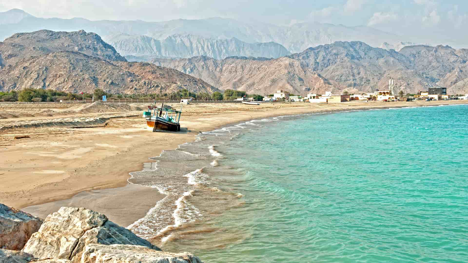 A serene scenic view of Sir Bani Yas Island, with a small traditional boat on a sandy beach and turquoise water, framed by rocky mountains in the background.