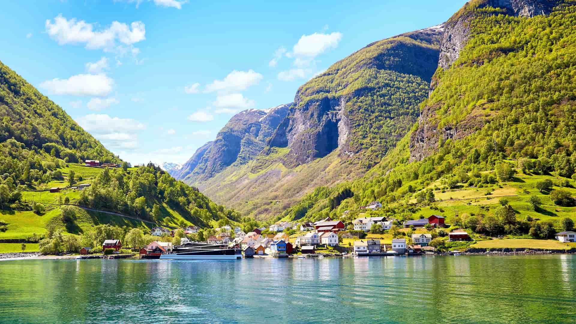 A breathtaking view of the Sognefjord in Norway, with a small village of colorful houses nestled in a valley between steep, lush green mountains, and the serene water reflecting the beautiful sky.
