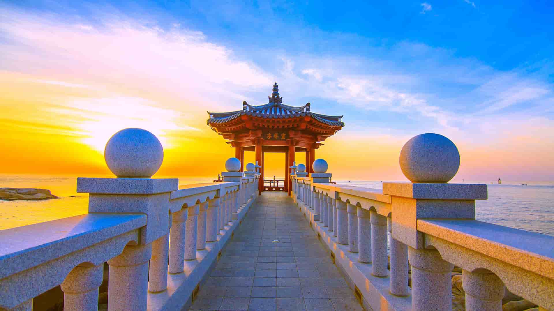 A scenic view of a traditional Korean pavilion at the end of a stone walkway, overlooking the East Sea in Sokcho, South Korea, with a stunning sunset in the background.