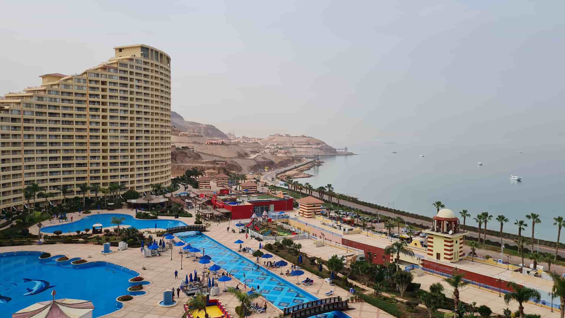 An expansive aerial shot of a luxury resort in Ain Sokhna, Egypt, with large swimming pools, water slides, and palm trees next to the Red Sea, and a mountainous landscape in the background.