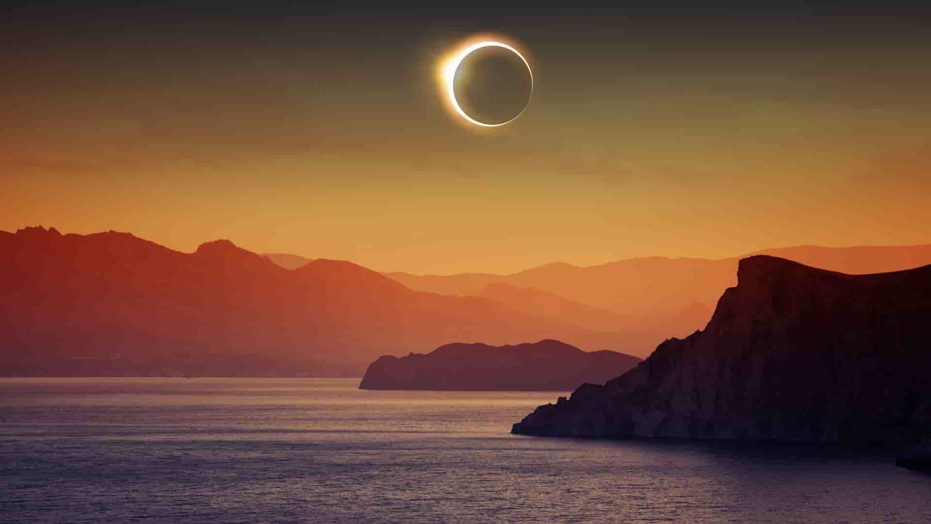 A dramatic celestial event over the sea with a ring of light, the total solar eclipse, visible over a tranquil body of water surrounded by silhouetted mountains.
