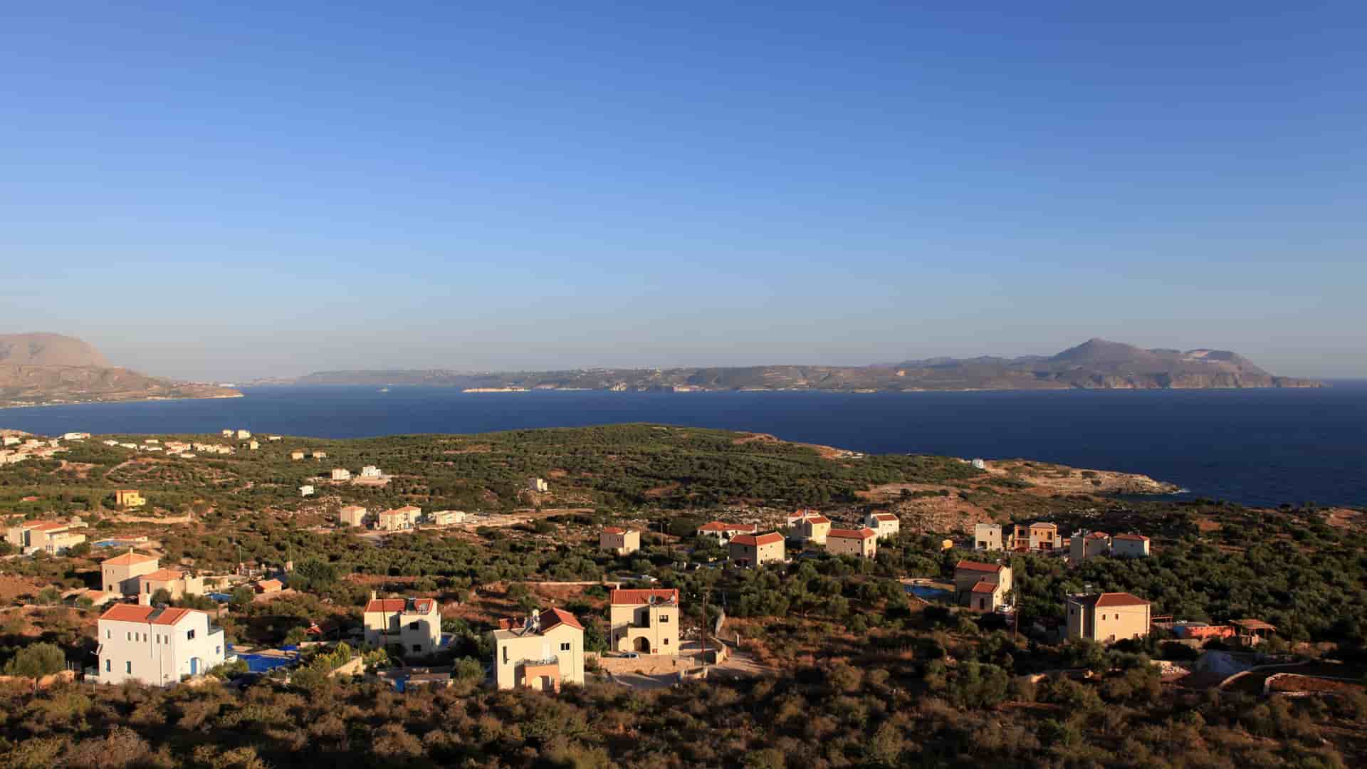 A high-angle view of a coastal village with low-rise buildings and olive groves on a hillside overlooking the tranquil waters of Souda Bay in Chania, Crete.