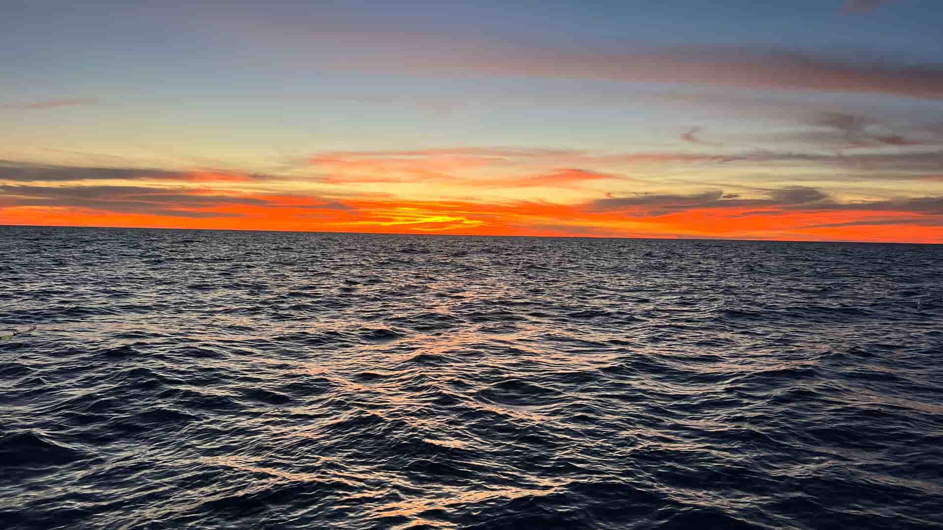 A wide-angle view of a fiery orange and red sunset over the deep blue, rippling waters of the South China Sea, with a straight horizon.
