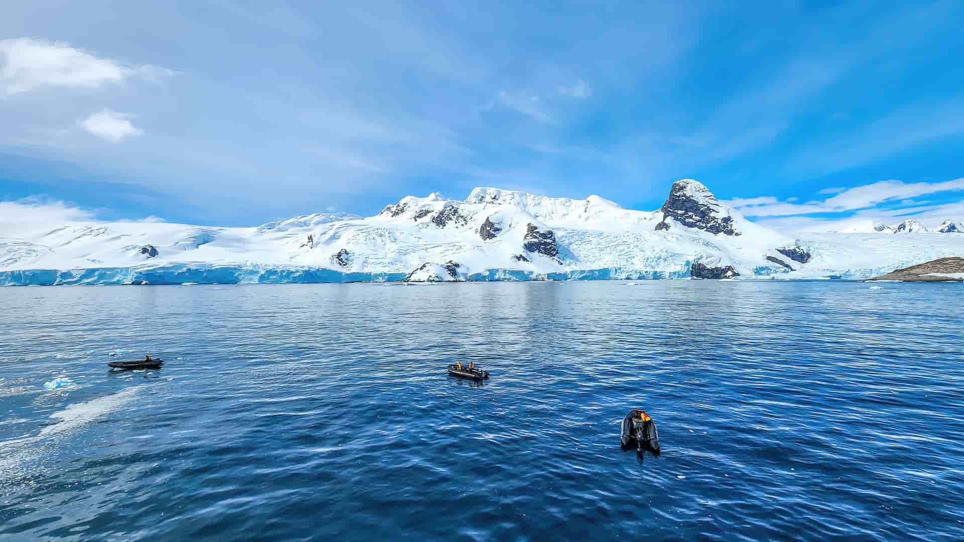 A stunning panoramic view of the South Shetland Islands, with a majestic snow-covered coastline and glaciers meeting the deep blue water, with a few small boats in the foreground.