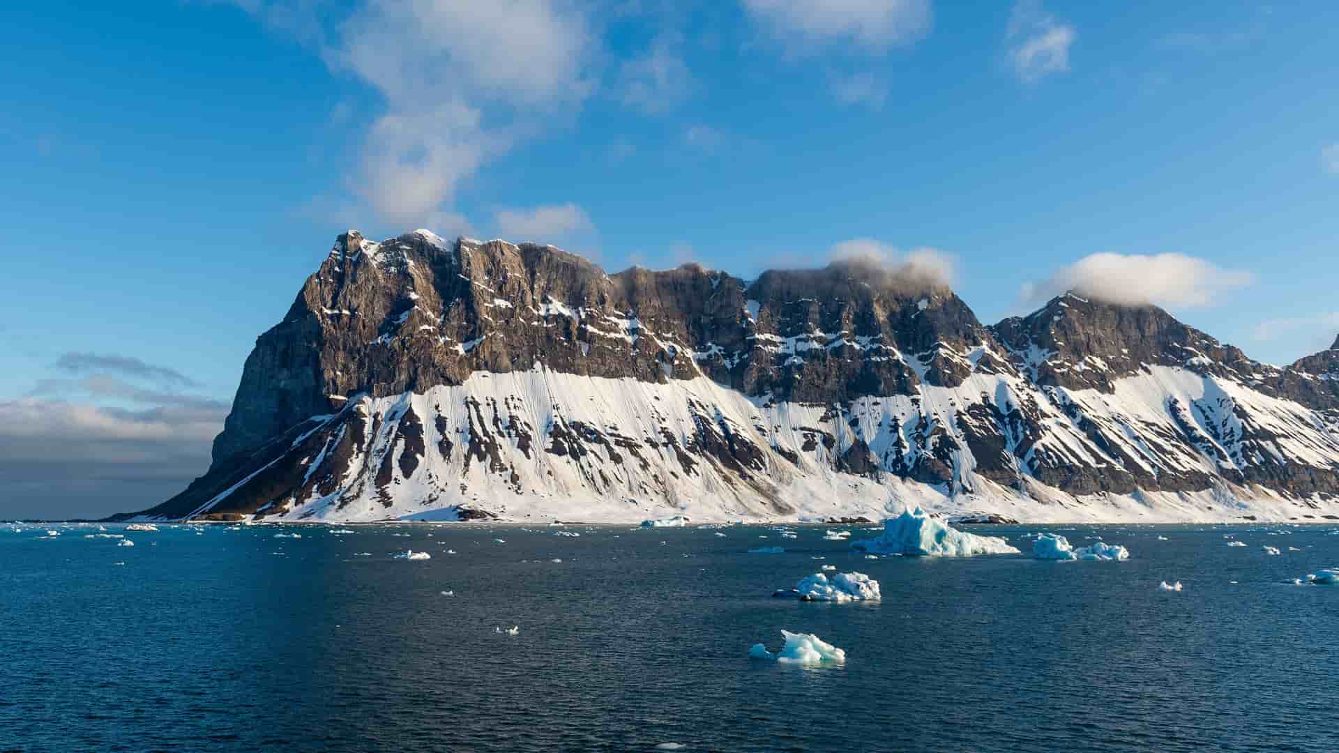 A panoramic view of the majestic, snow-covered mountains of Southern Svalbard, Norway, with small icebergs and glaciers floating in the deep blue sea.