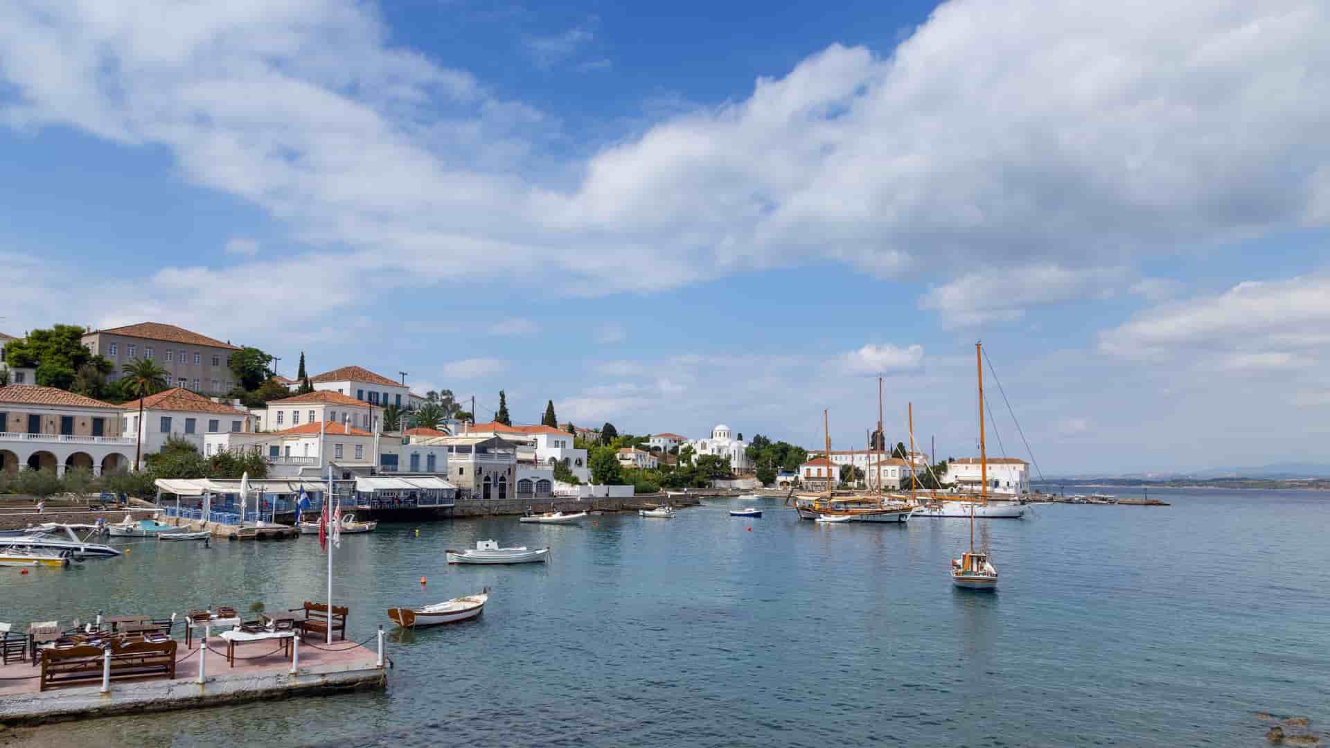A charming view of the old harbor in Spetses, Greece, with traditional white buildings and classic wooden sailboats moored in the calm bay under a bright sky with scattered clouds.