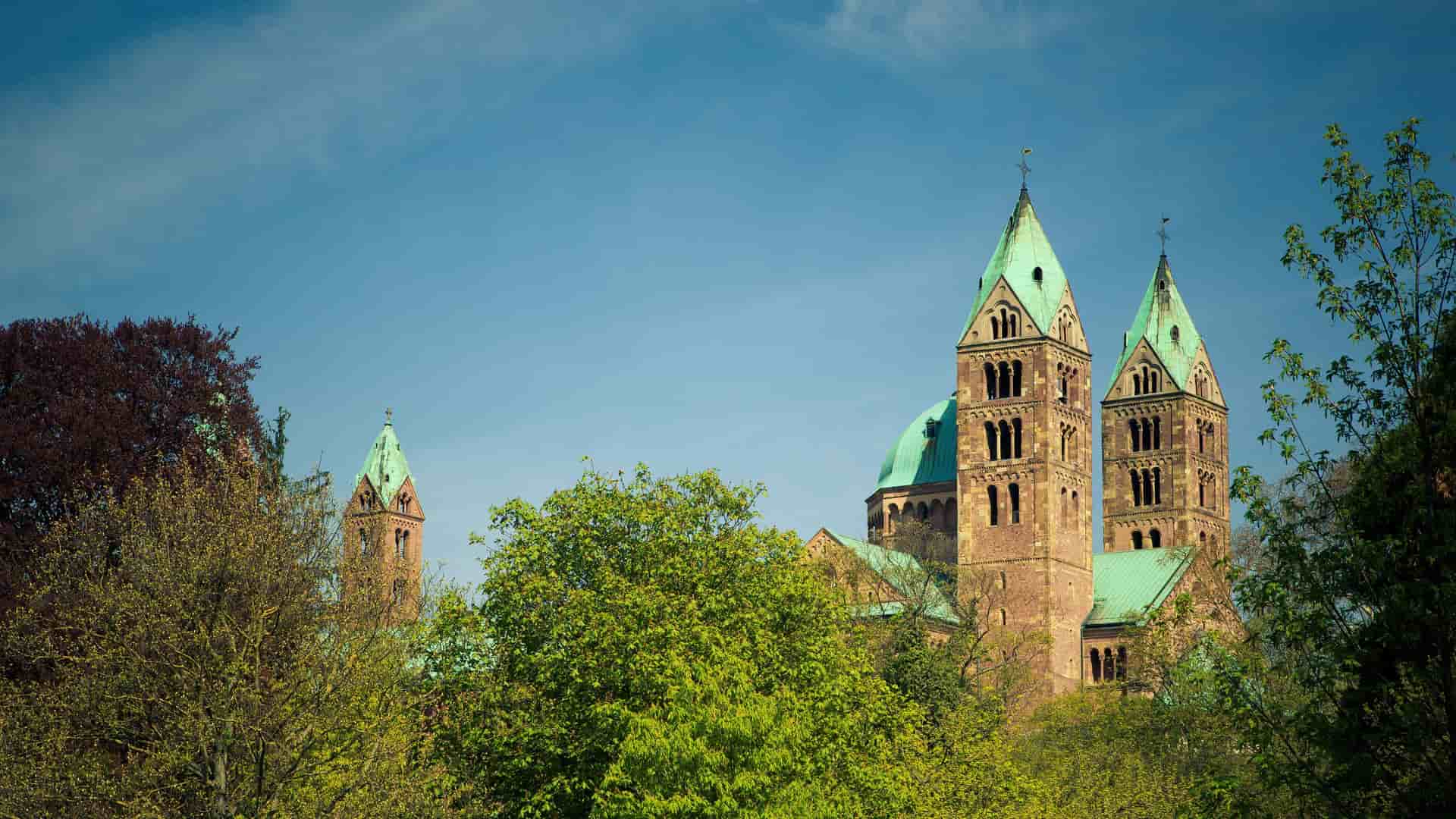 A beautiful view of the Speyer Cathedral with its iconic twin green-roofed towers peeking above the lush green trees against a clear blue sky in Speyer, Germany.
