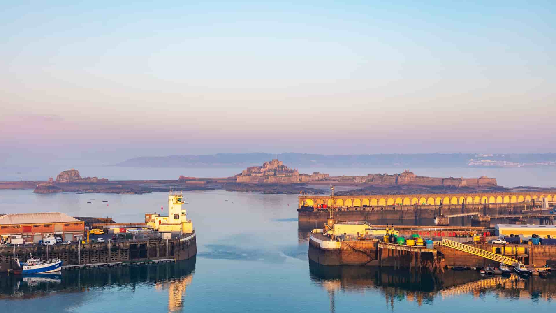 A panoramic view of the harbor in St. Helier, Channel Islands, with a tranquil bay, a port with docked boats in the foreground, and the historic Elizabeth Castle in the background under a beautiful morning sky.