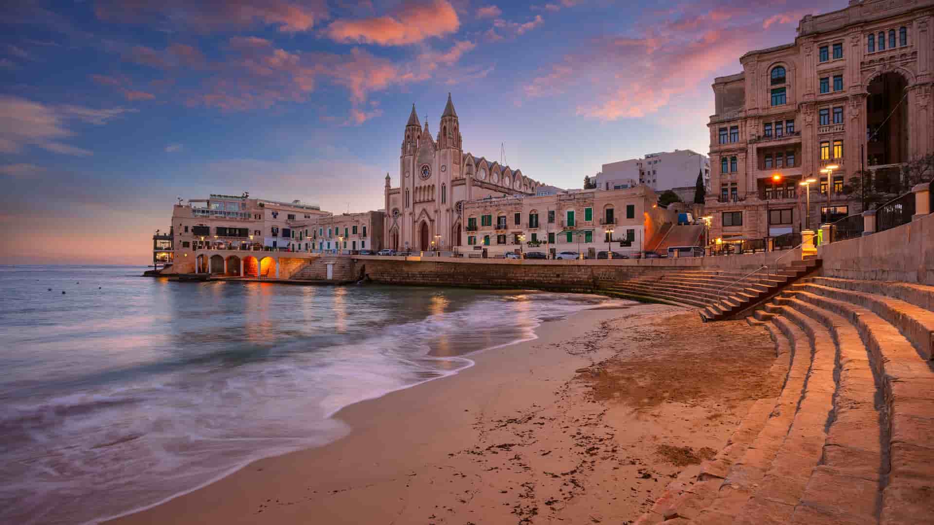 "A breathtaking sunset view of St. Julian's, Malta, with a sandy beach and gentle waves, showcasing the iconic Parish Church of Our Lady of Mount Carmel and the illuminated town buildings in the background.  "