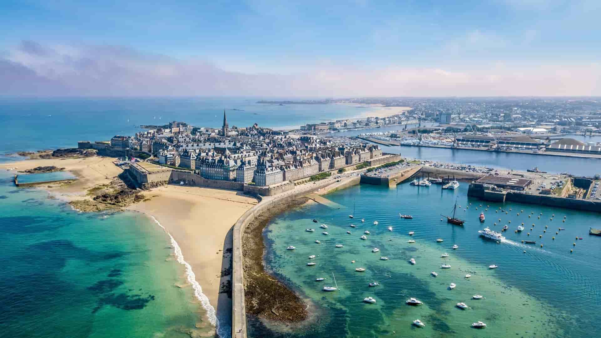 An aerial view of the historic walled city of St-Malo, France, with its medieval buildings and walls, a sandy beach, and a harbor full of sailboats and boats in the foreground.