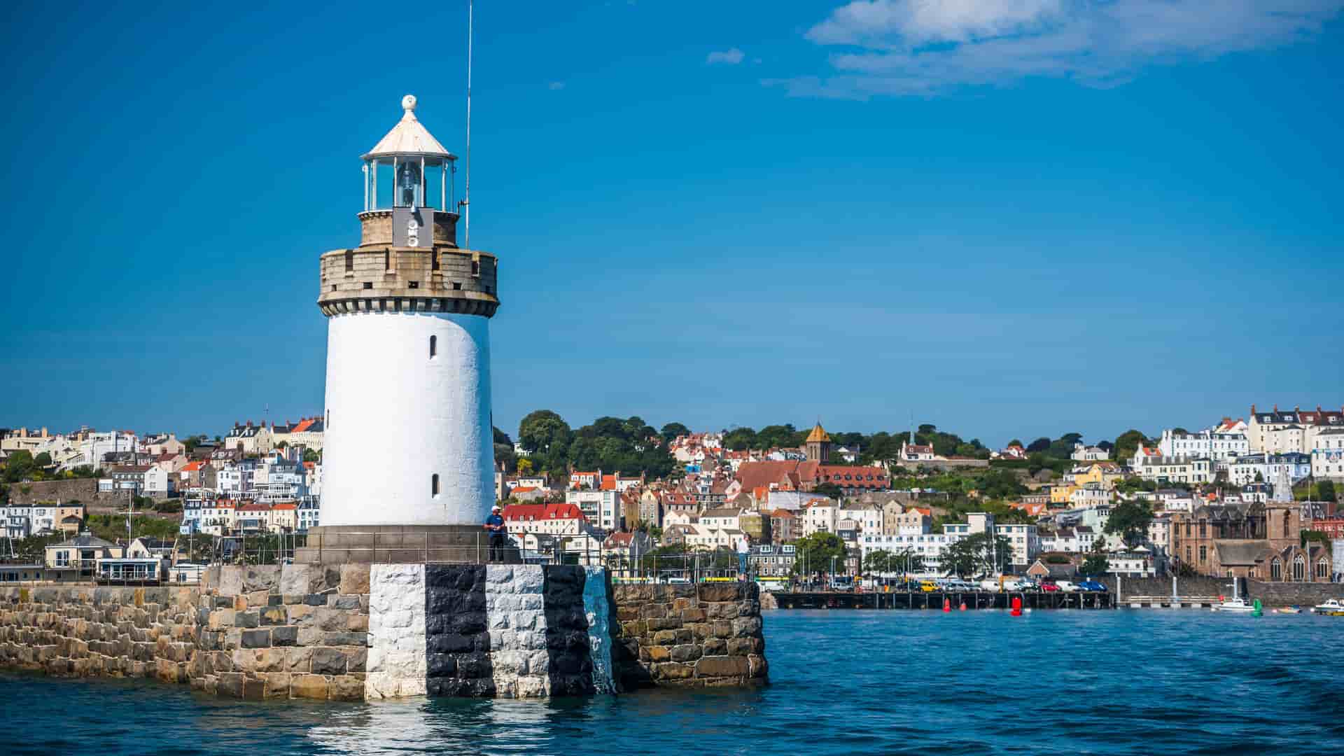A view of St Peter Port, Guernsey, with its distinctive black-and-white-striped lighthouse on a stone jetty in the foreground and the colorful town and harbor in the background.