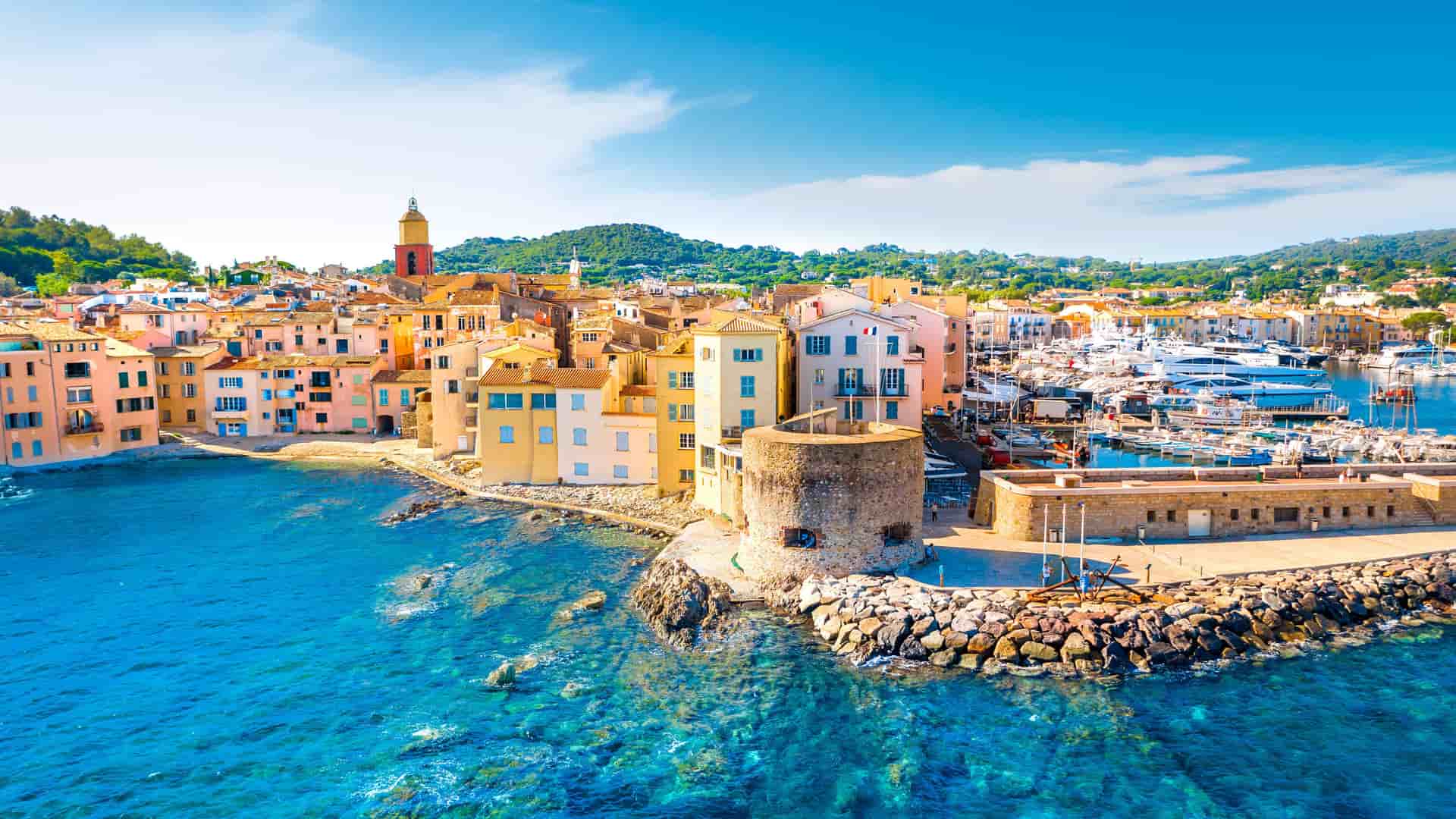 A wide-angle aerial view of the iconic harbor of St. Tropez, France, with its vibrant port full of luxurious yachts and sailboats, and the colorful buildings along the waterfront.