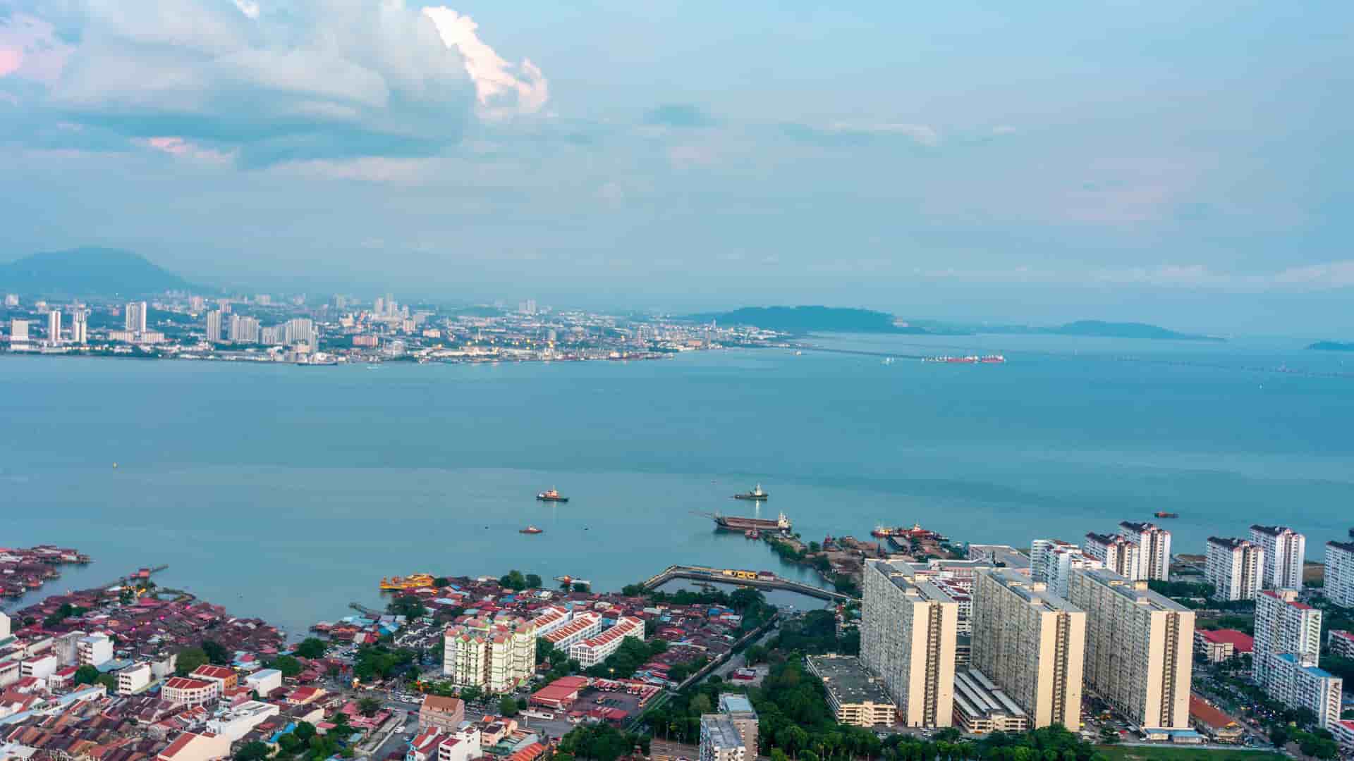 An aerial view of the bustling city of George Town, Malaysia, on the coast of the Strait of Malacca, with ships in the water and modern buildings surrounded by lush green mountains.
