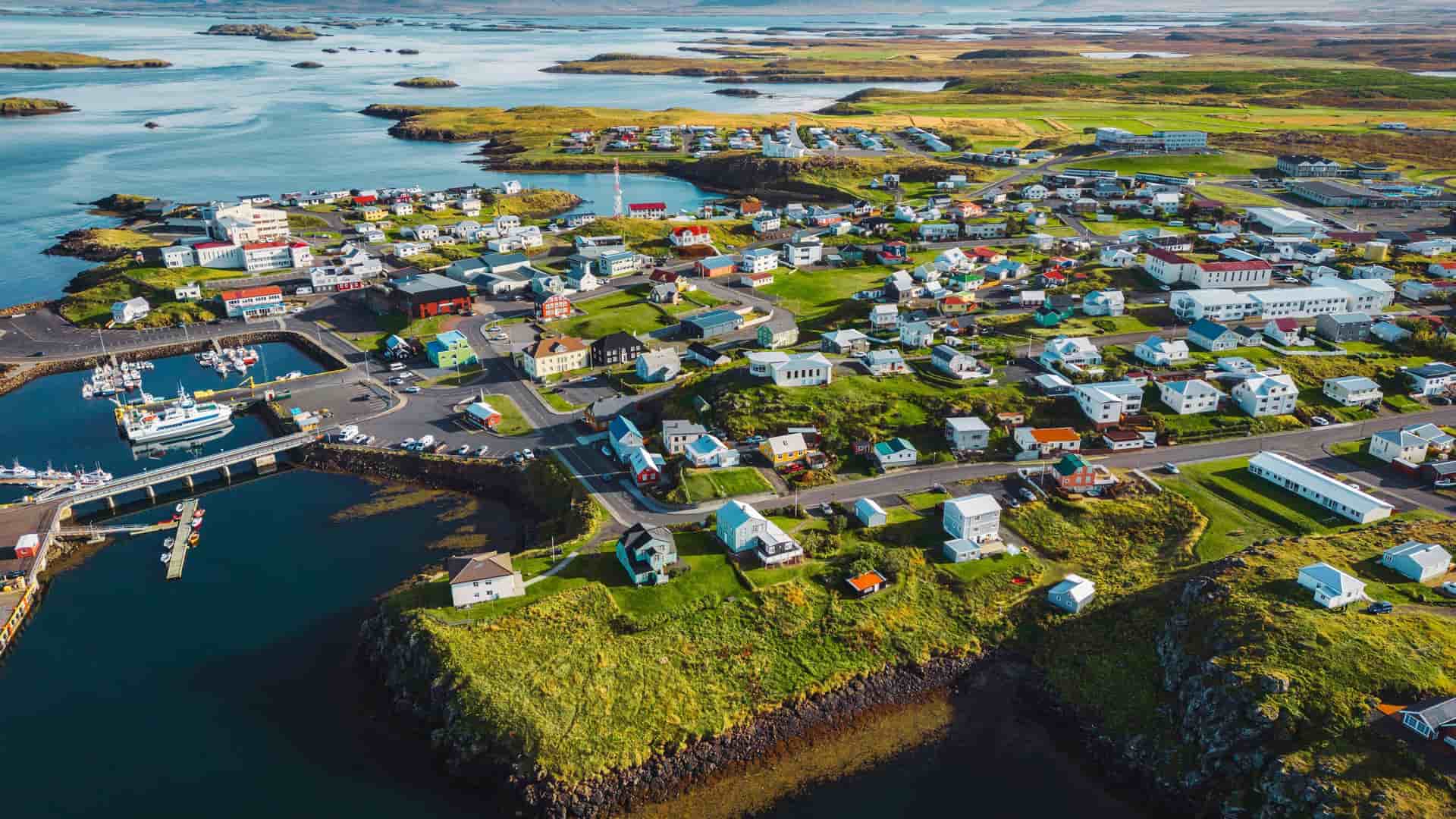 An aerial view of the vibrant fishing town of Stykkishólmur, Iceland, showing colorful houses, a harbor with boats, and the surrounding green landscape dotted with small islands.