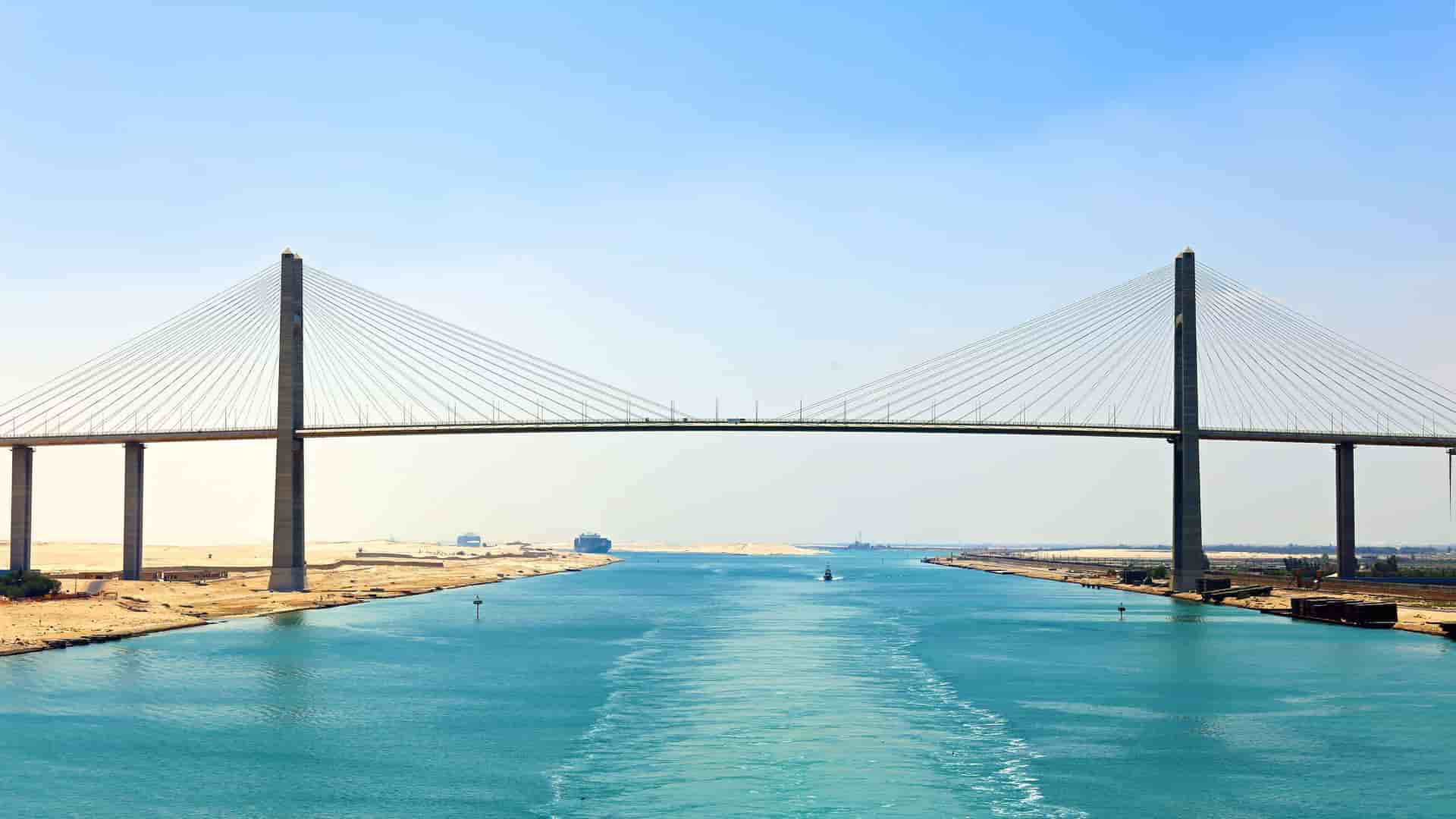 A massive suspension bridge, the El Ferdan Railway Bridge, spanning the Suez Canal in Egypt, with a large container ship visible in the distance.