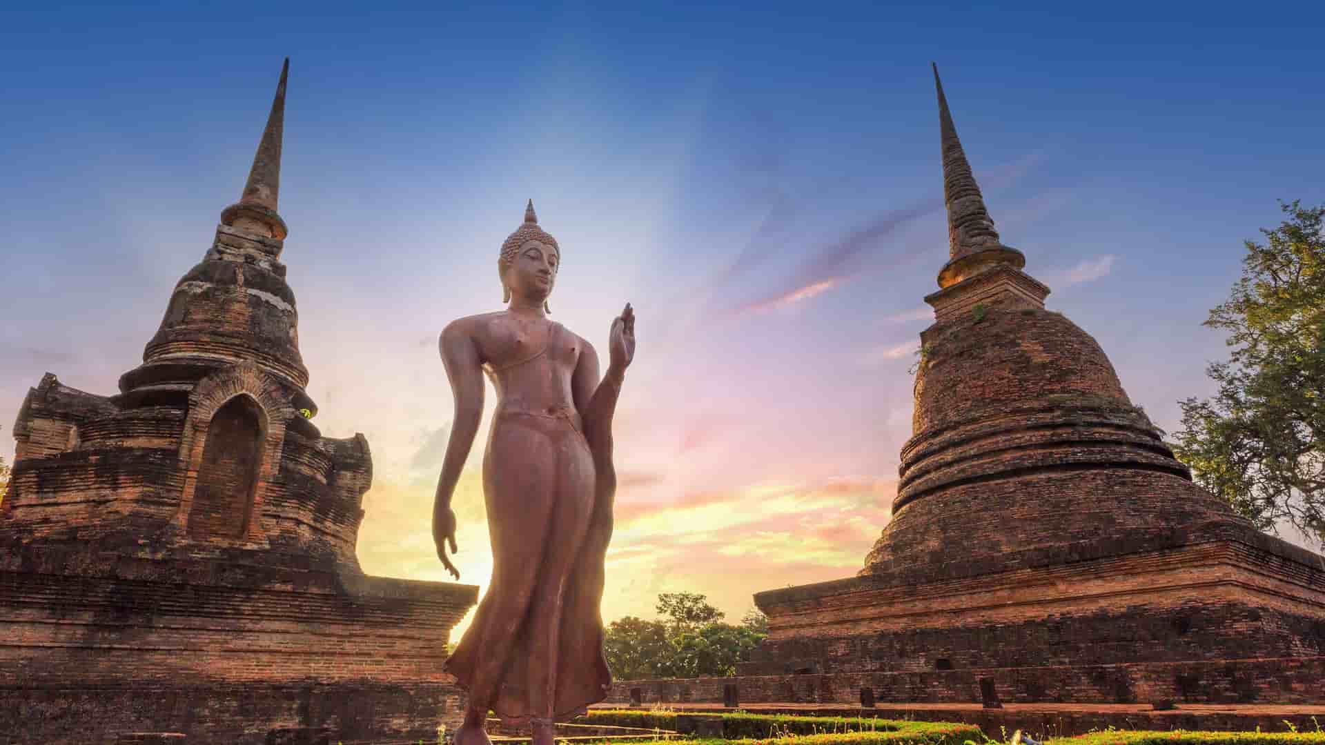 "A towering bronze Buddha statue with a distinctive walking pose, standing between two ancient brick temple ruins at the historic Sukhothai Historical Park in Thailand.  "