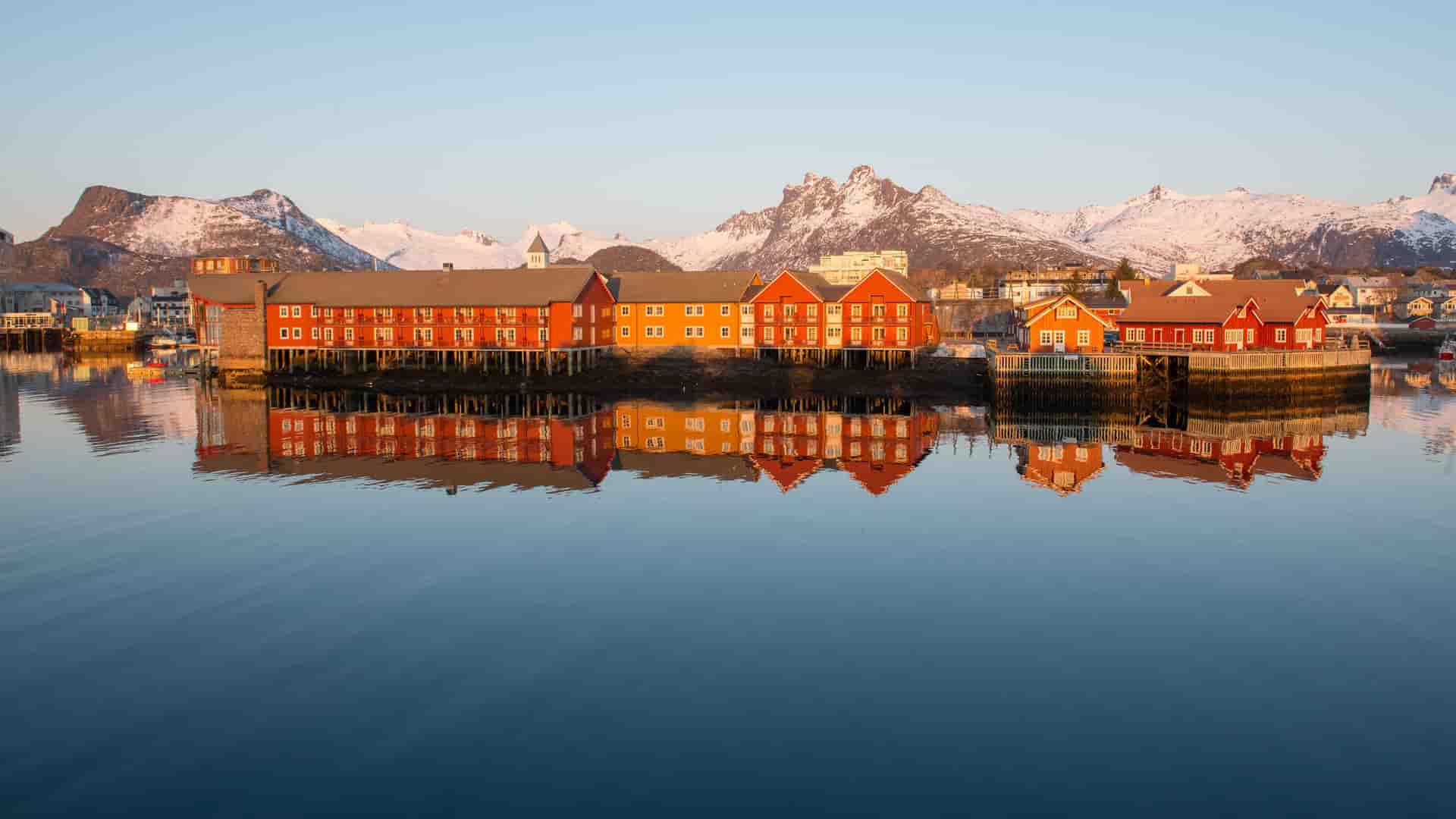 A serene panoramic shot of the Svolvær harbor in Norway, with traditional red and orange fishing cabins, or rorbu, reflected in the calm water and a backdrop of majestic snow-capped mountains.