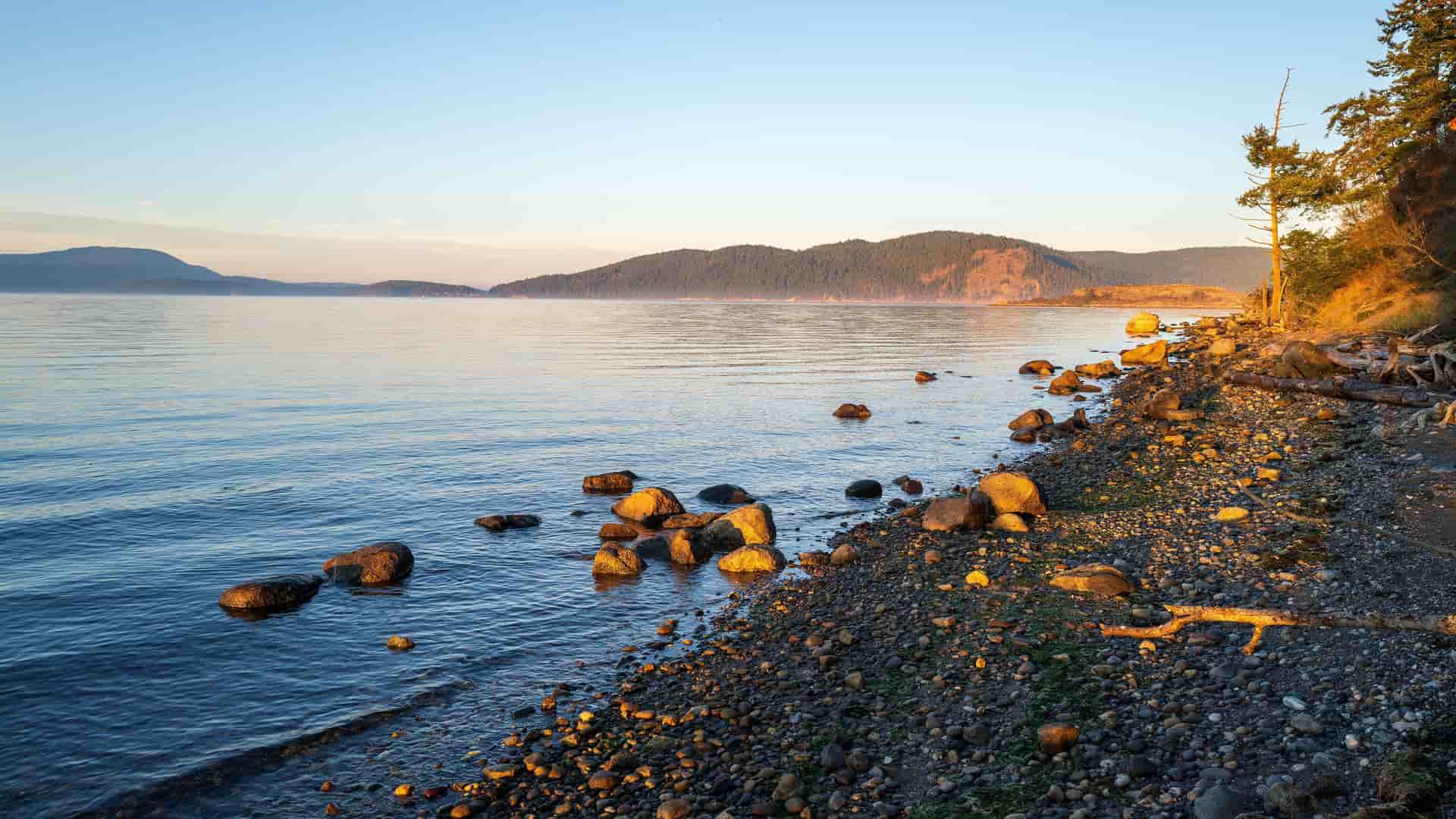 A serene rocky beach at Swift Bay in Alaska, with the calm waters of the bay reflecting the soft light of a golden sunset.