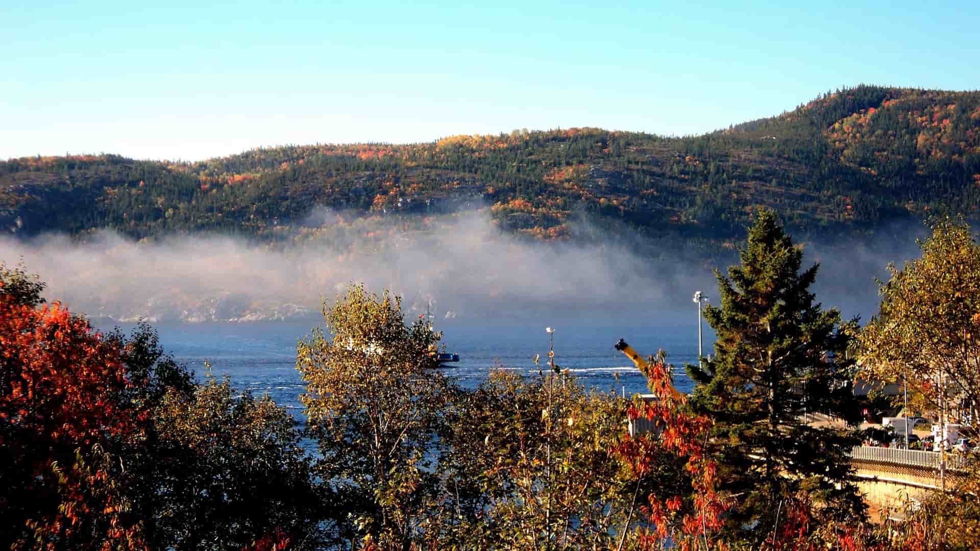 A beautiful autumn landscape of the Saguenay Fjord in Tadoussac, Quebec, with colorful fall foliage and mist rising from the water.