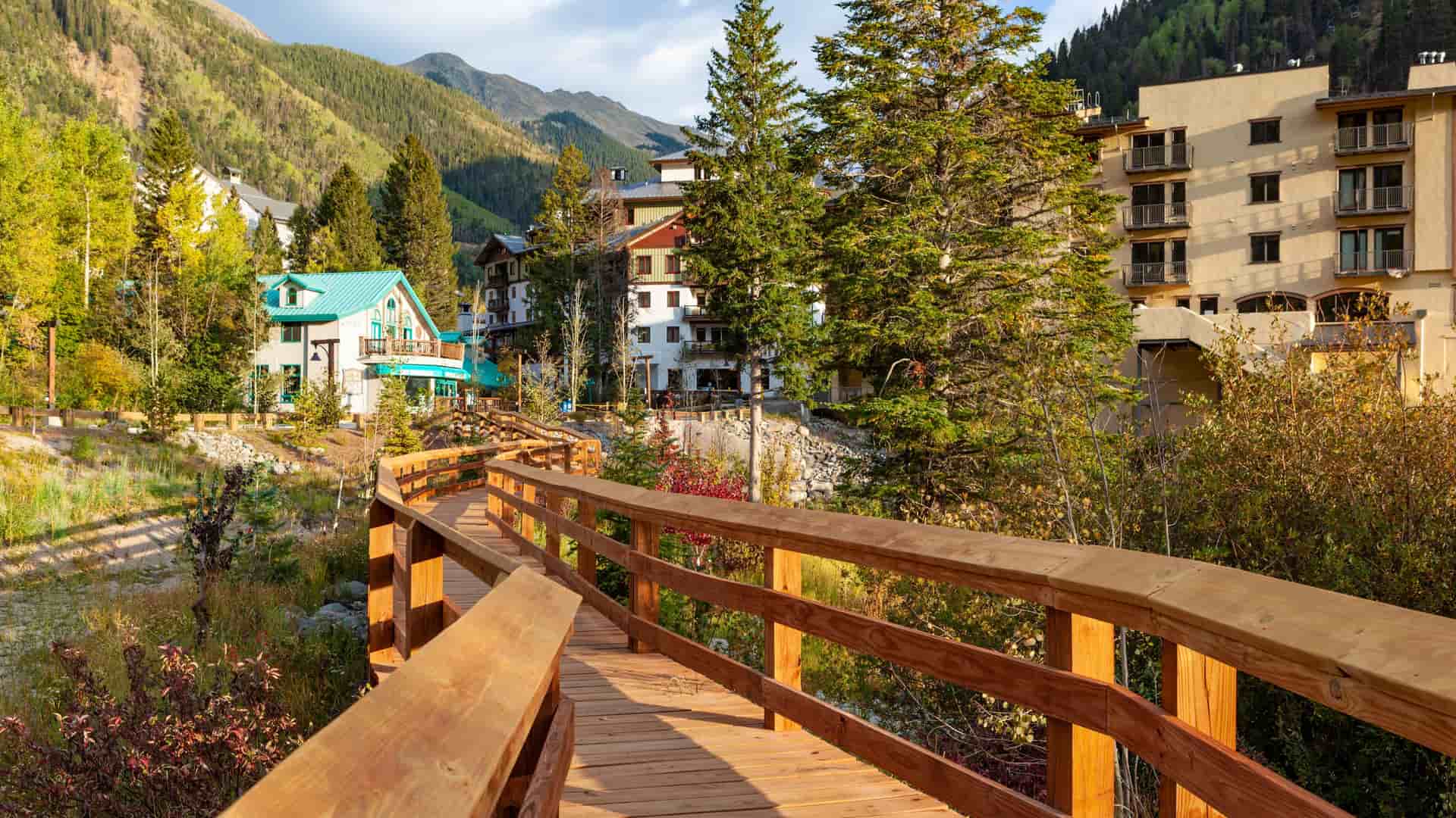 "A rustic wooden walkway winds through the colorful autumn foliage toward the alpine ski resort village buildings nestled in the mountains of Taos, New Mexico.  "
