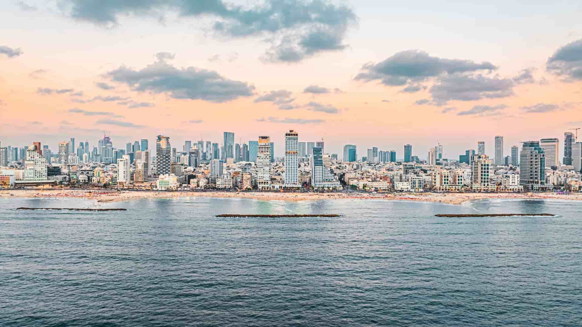 "An aerial view of the city of Telavi, Georgia, showing the dense urban landscape of buildings with red-tiled roofs surrounded by lush green trees and fields, with the Caucasus Mountains in the distant background.  "