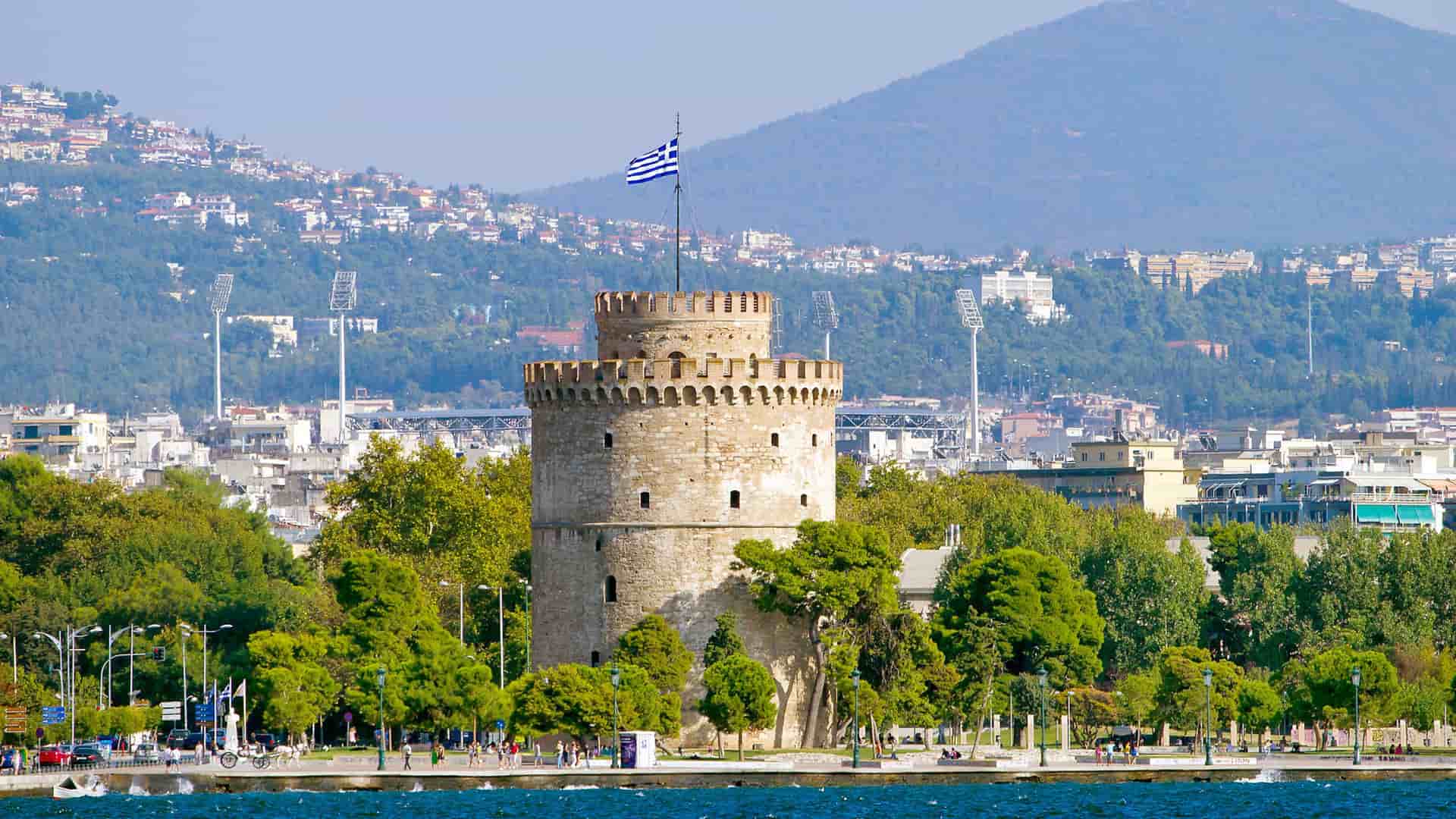 A striking view of the historic White Tower of Thessaloniki, a major landmark on the waterfront, with the city's buildings and surrounding hills in the background.