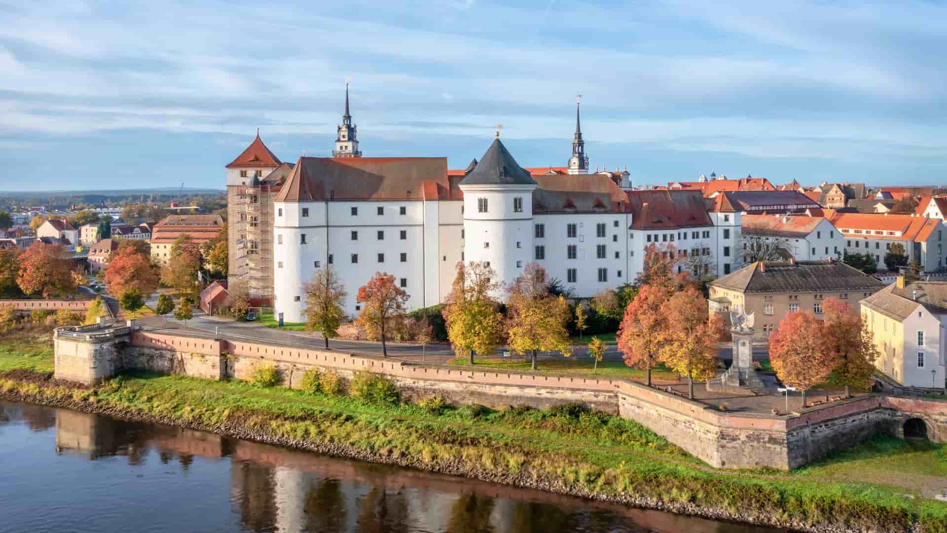 A picturesque view of Hartenfels Castle, a major landmark in Torgau, Germany, with its Renaissance architecture and towers along the Elbe River, surrounded by trees with vibrant autumn foliage.