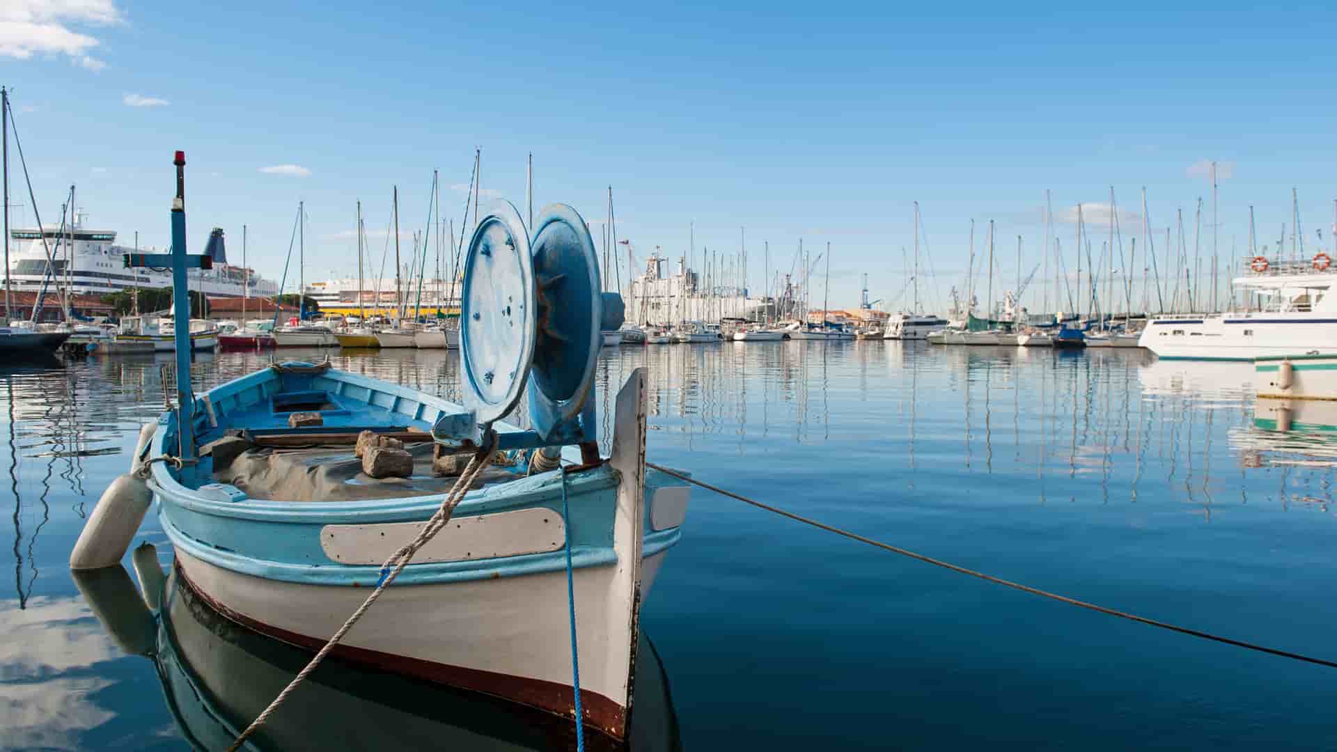 A classic blue fishing boat docked in the busy port of Toulon, France, with numerous sailboats and yachts reflected in the calm water.