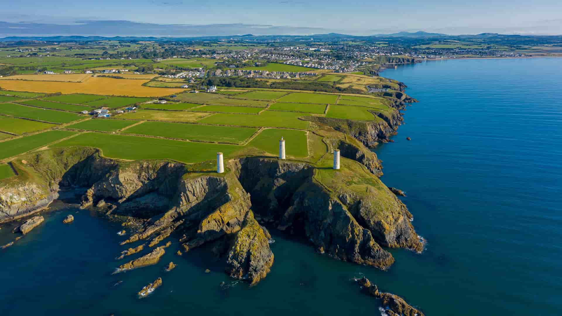 "An aerial view of the rugged coastline of Tramore, Ireland, showcasing the three historic stone pillars standing on the cliff's edge, overlooking the deep blue sea. The surrounding landscape features a patchwork of green and yellow fields leading to a small town in the distance.  "